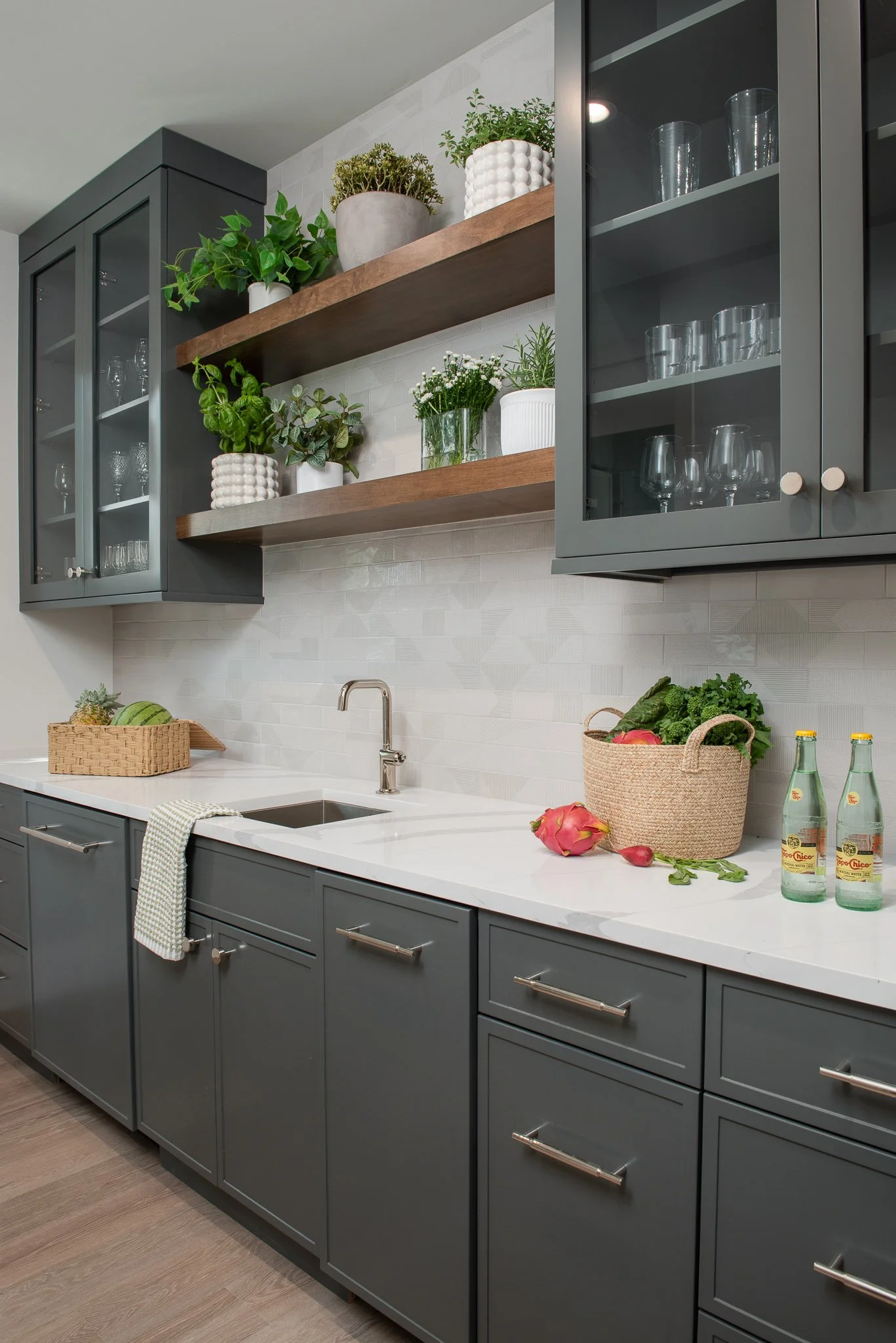 Modern kitchen with gray cabinets, white countertop, open wooden shelves with potted plants, and glass-front cabinets with glassware.