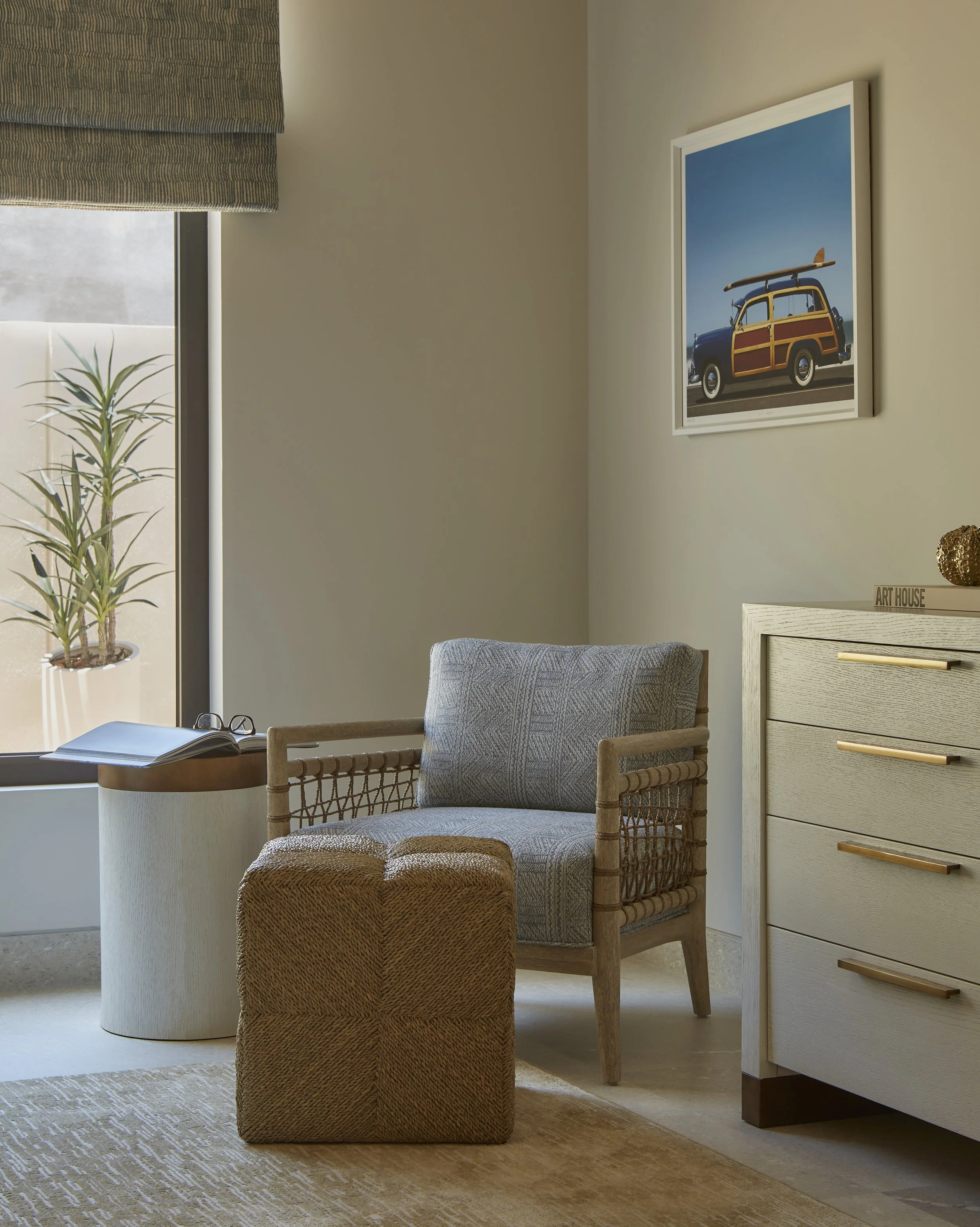 Living room with a beige armchair, woven ottoman, side table with open book and glasses, potted plant near window, framed picture of a wood-paneled car with surfboard on wall, beige dresser with decorative items.