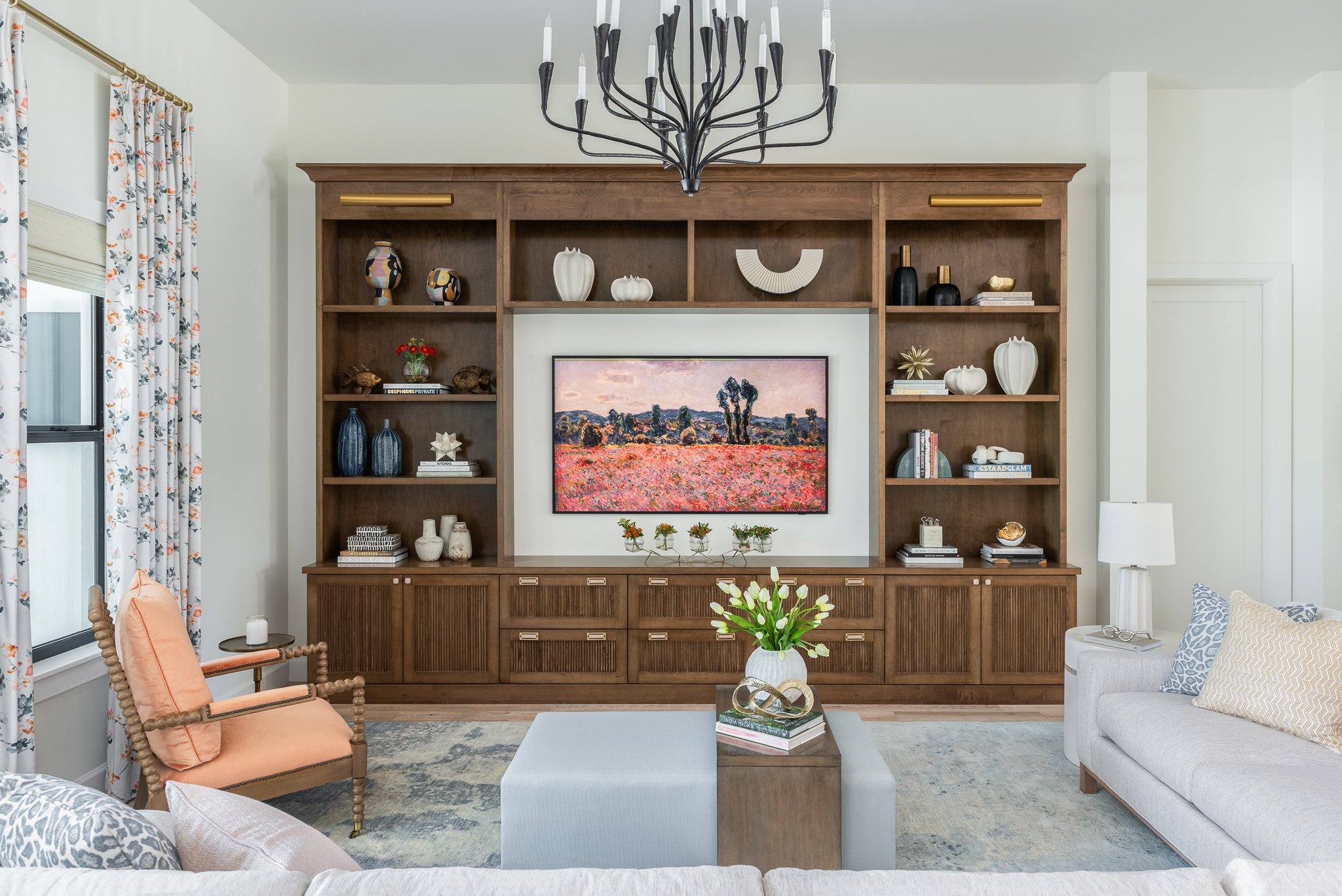 Living room with built-in wooden shelves, a large framed landscape painting, a chandelier, floral curtains, a peach armchair, a white sectional sofa, a gray ottoman, a side table with a lamp, and decorative items including vases, books, and flowers.