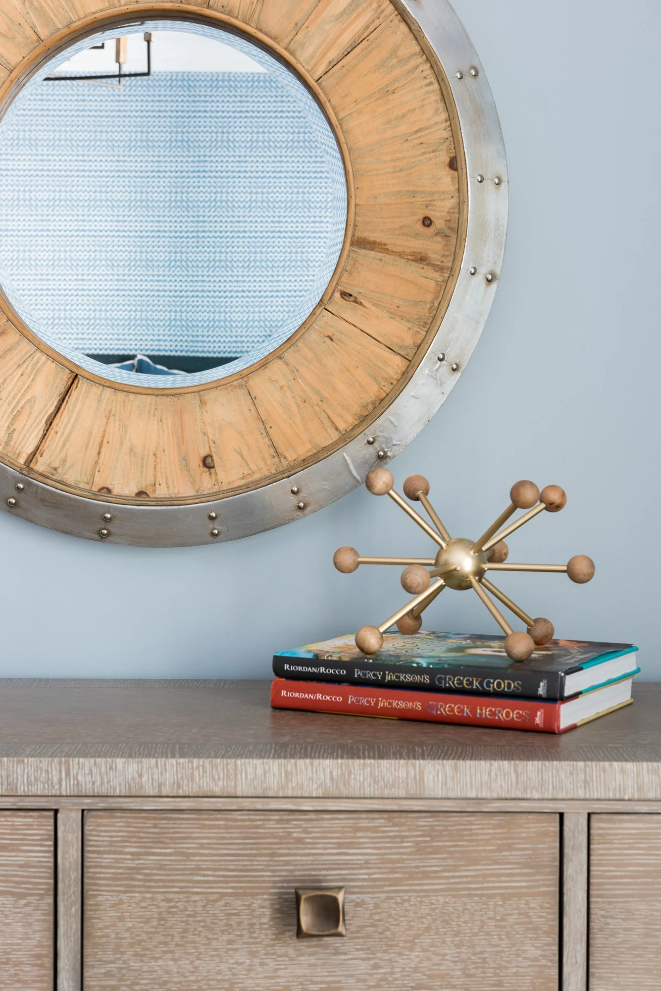 A round mirror with a wooden and metal frame hangs on a blue wall. On a wooden surface below, there are two stacked books about Greek mythology, and a decorative metallic atom sculpture.
