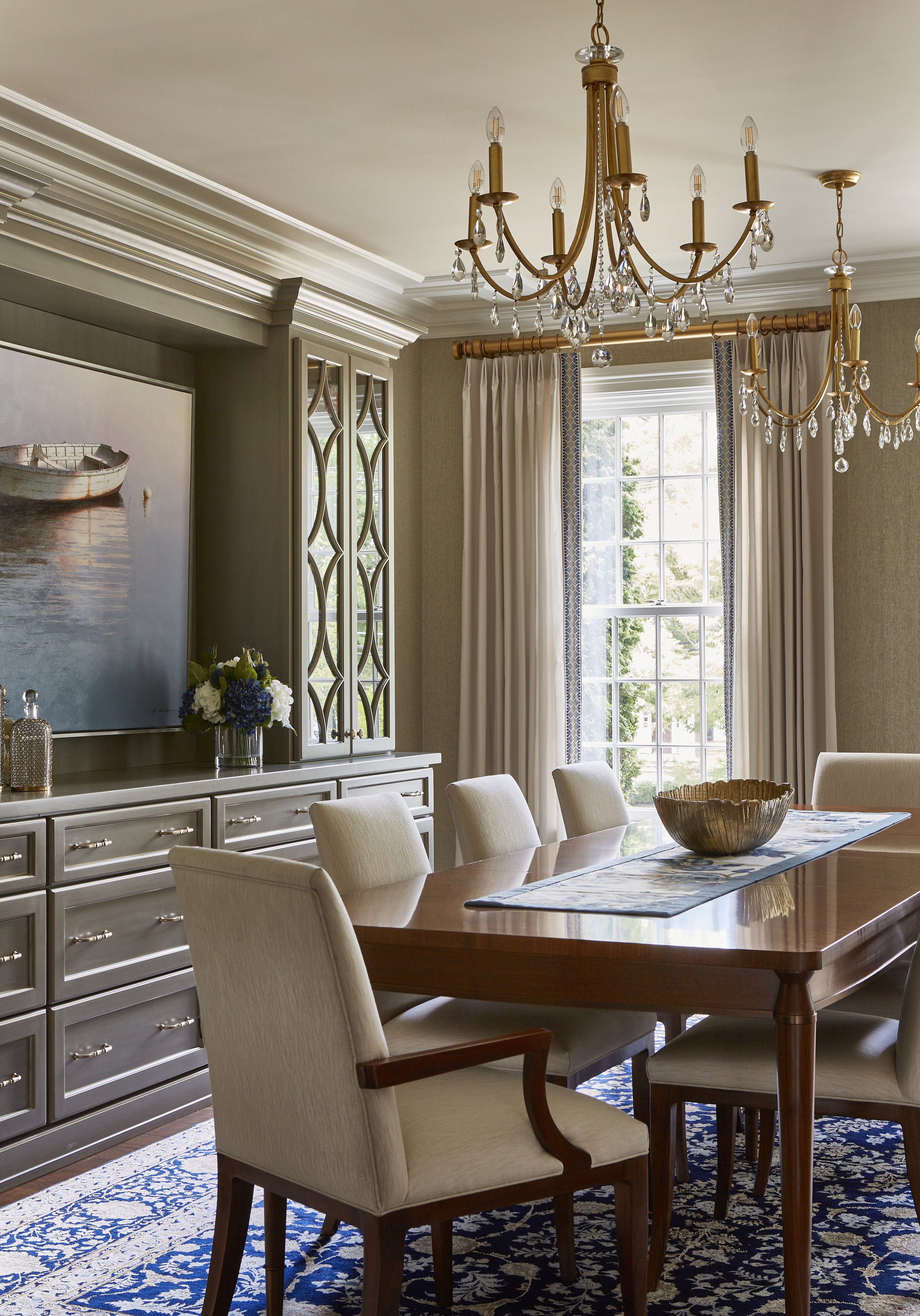 A dining room with a wooden table, beige upholstered chairs, a chandelier with crystals, a large window with curtains, a sideboard with a flower arrangement, and a patterned blue rug.