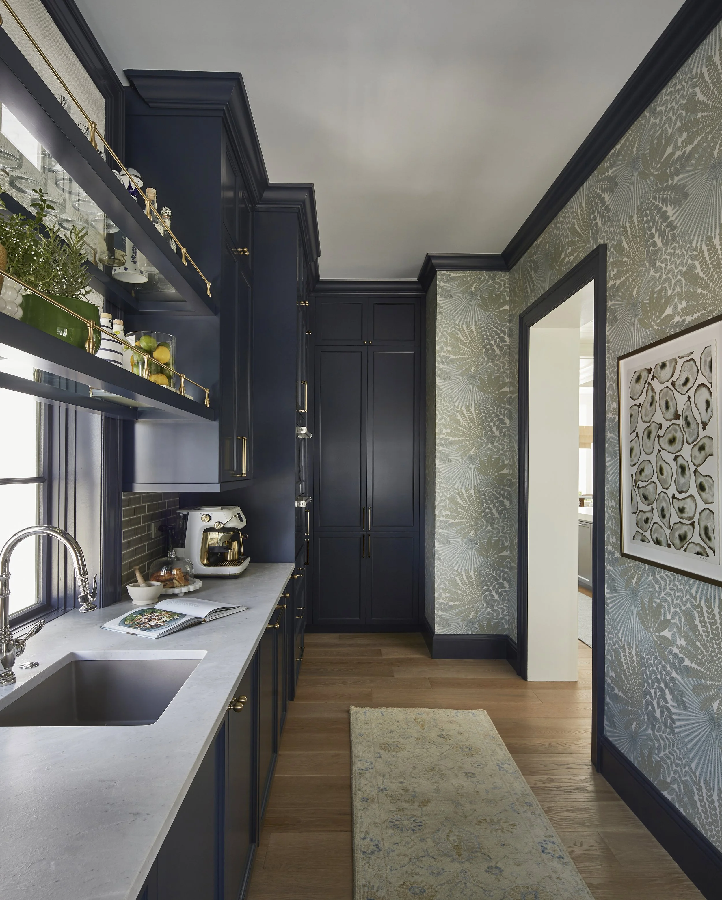Kitchen with navy blue cabinets, marble countertop, open shelves with glassware and plants, wooden floor, and wallpaper with green leaf patterns.