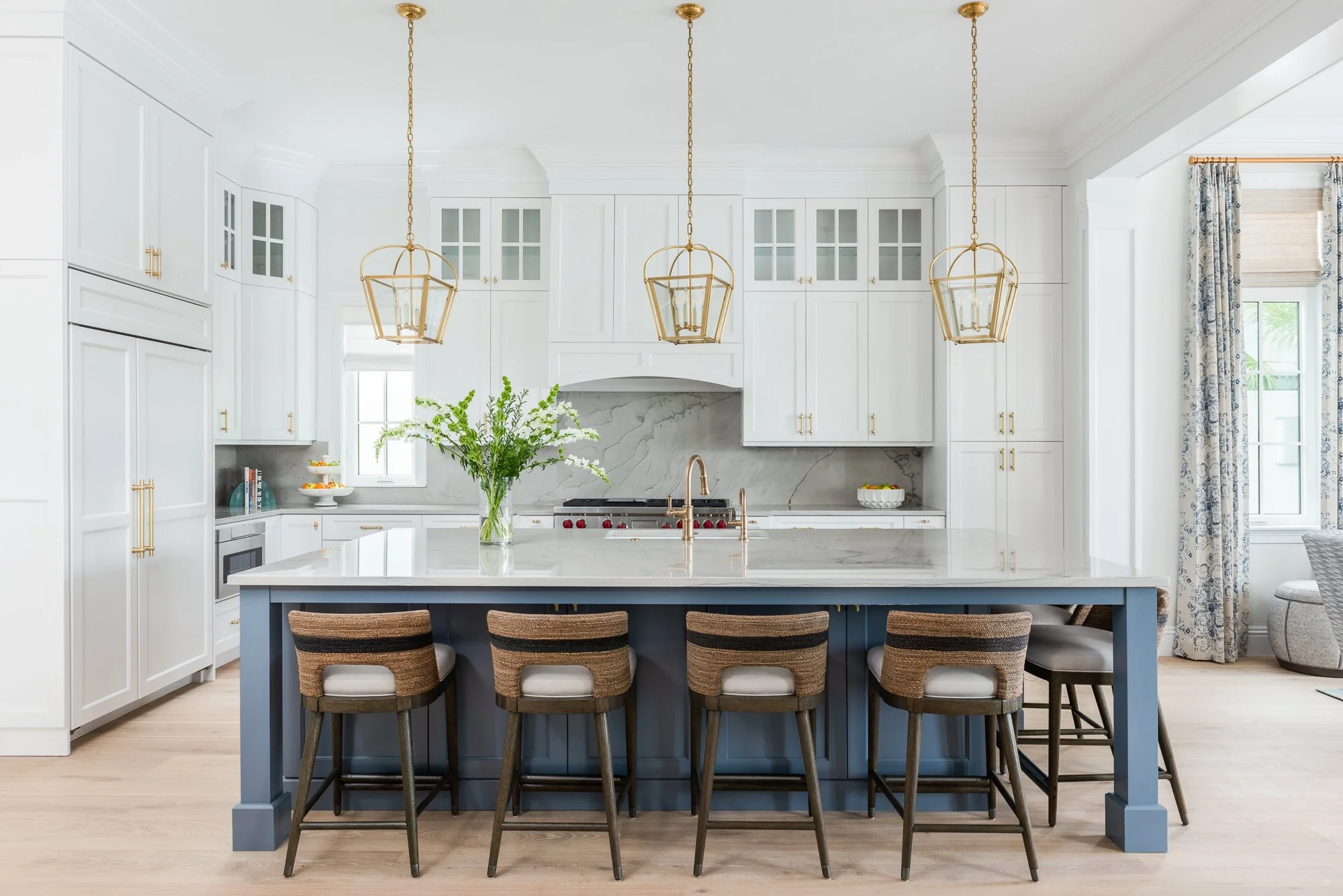 Modern white kitchen with a large blue island, gold pendant lights, a potted plant, and window with curtains.