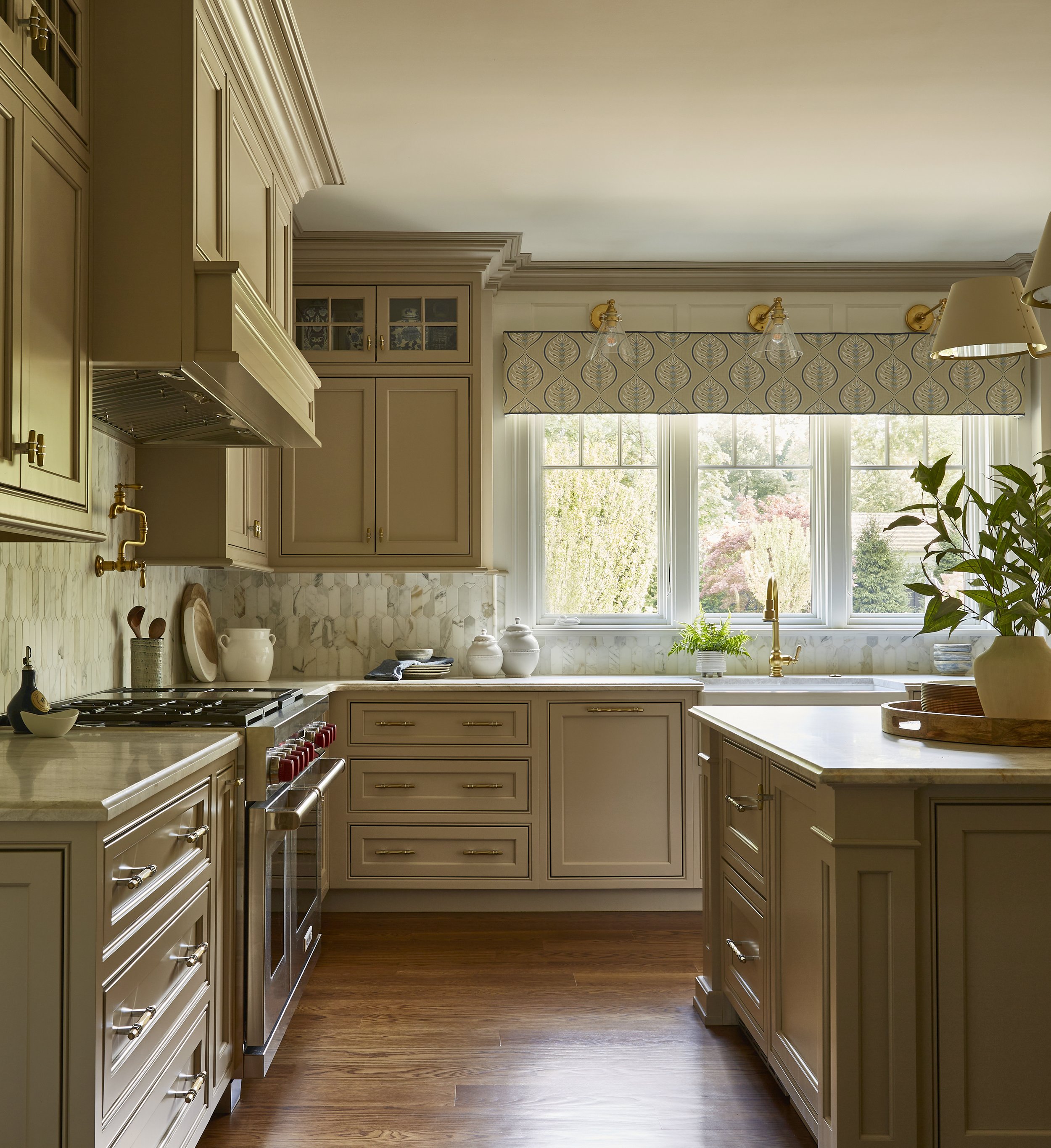 A bright kitchen with cream-colored cabinets, a marble backsplash, wooden flooring, and a large window with a patterned valance, overlooking a garden.
