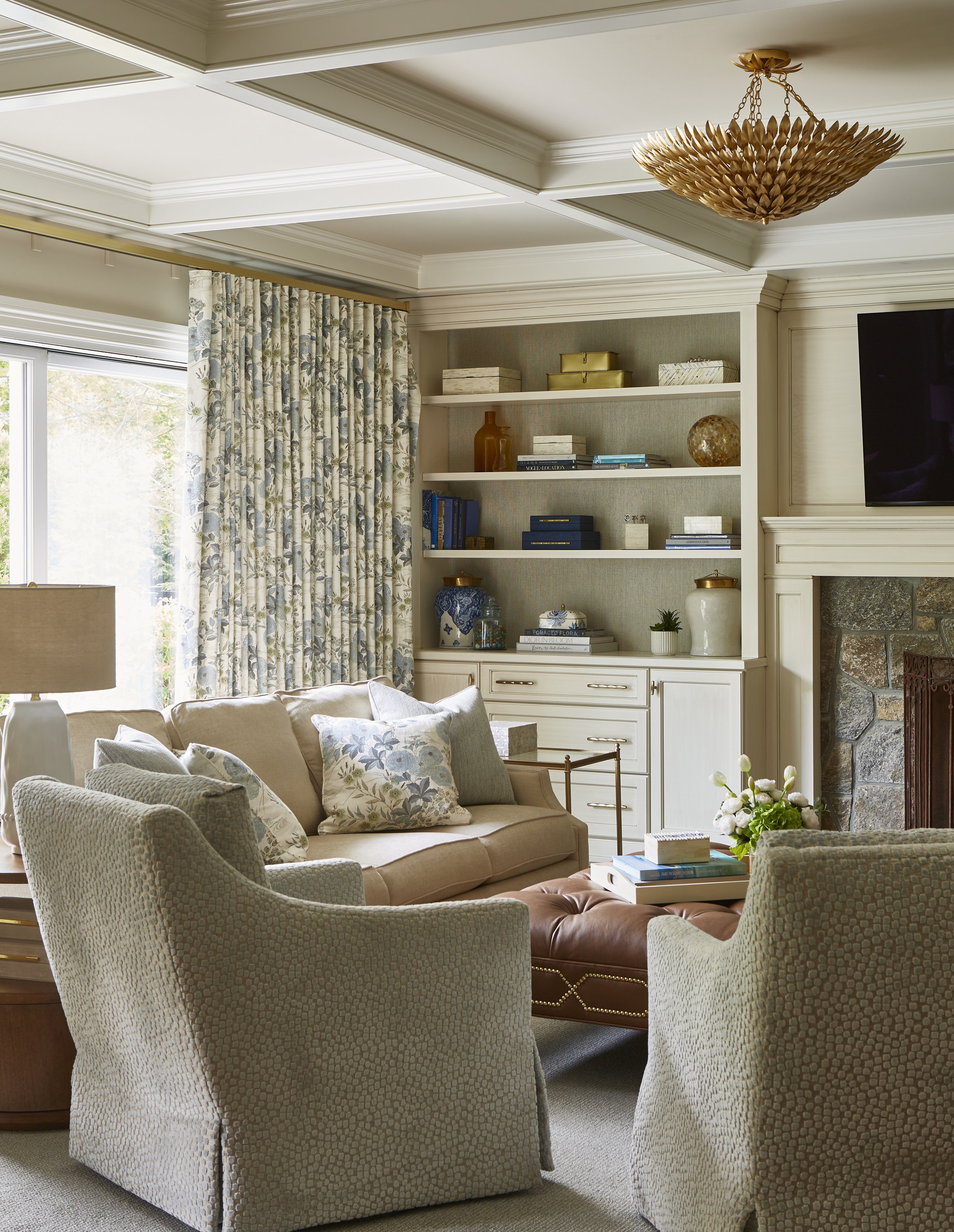 Living room with beige sofa, two patterned armchairs, a leather ottoman with a stack of books, a white built-in bookshelf with decorative objects and books, a stone fireplace, and a flat-screen TV on the wall. There are floral curtains by the window 