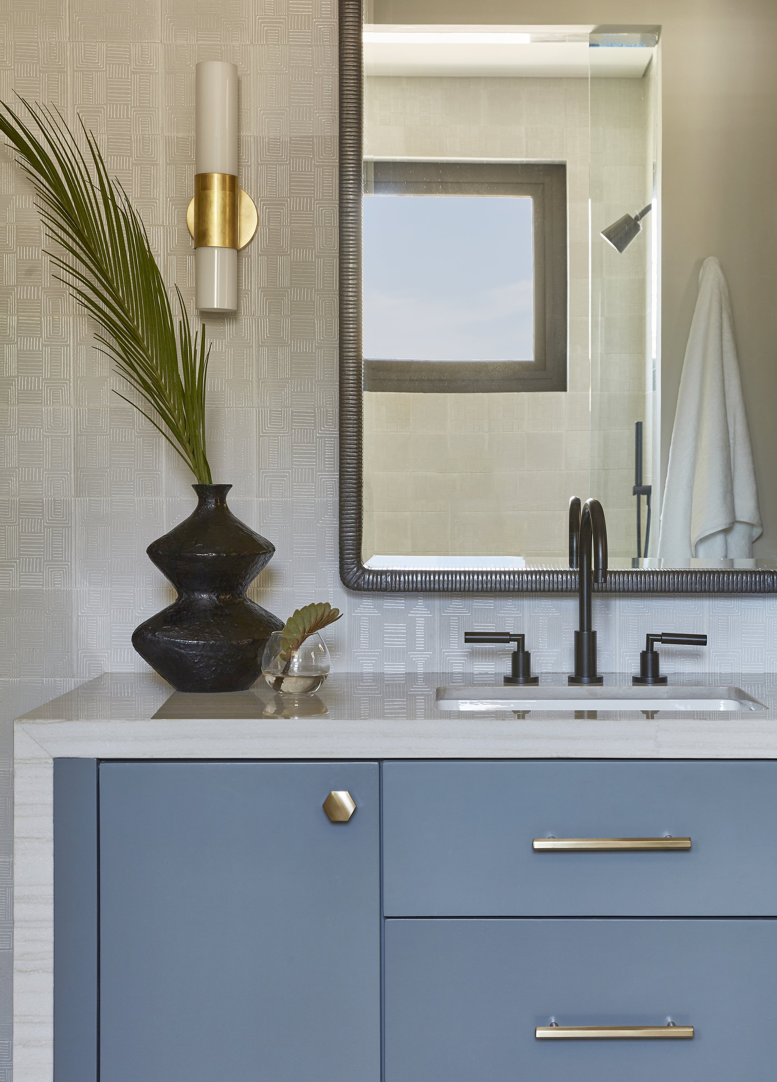 A bathroom with a blue vanity, a white countertop, and black fixtures. A large mirror reflects a window and a shower with a white towel. Decorative items include a black vase with green leaves and a small glass bowl with a leaf. There is a wall-mount