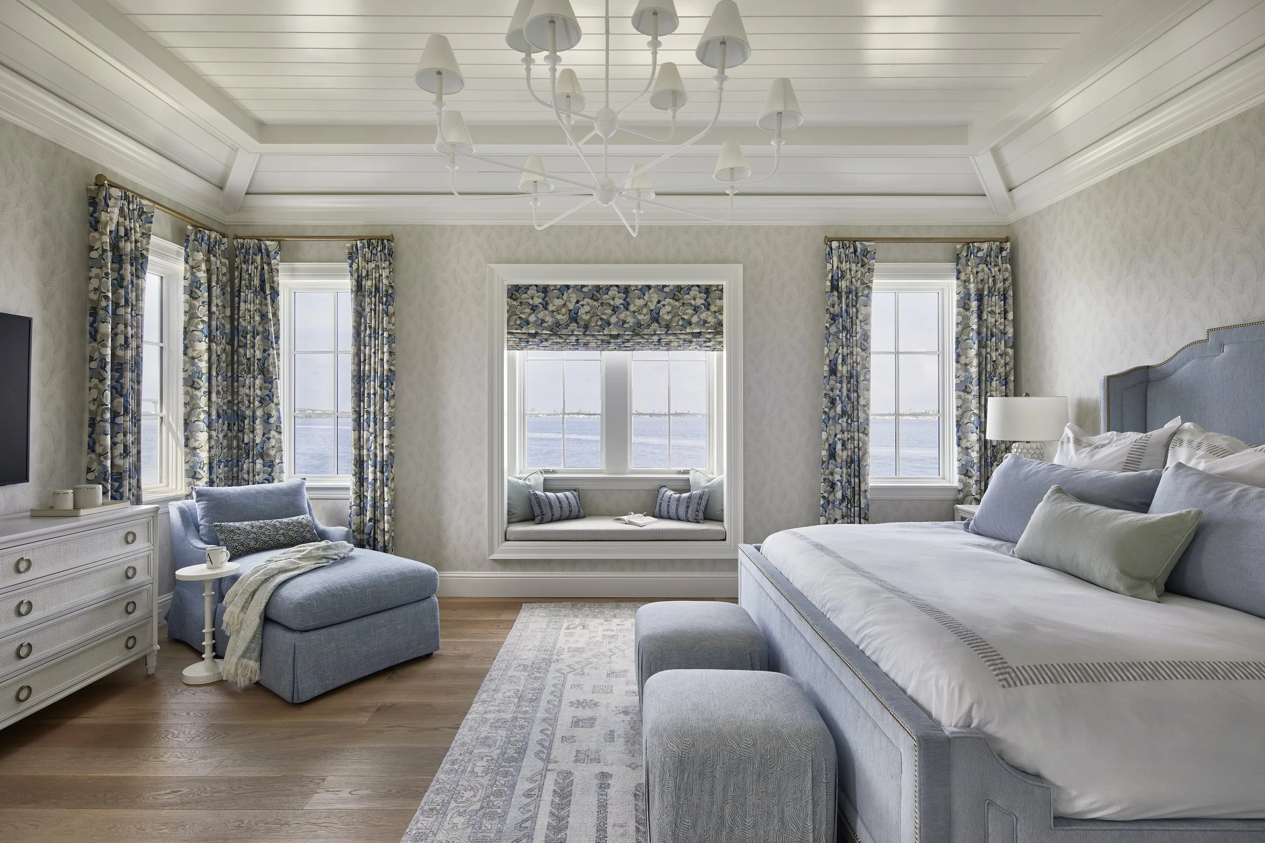 A bedroom with white walls, three large windows with floral curtains, a blue upholstered bed, a sitting area with a blue armchair, a white dresser, and a window seat overlooking water.
