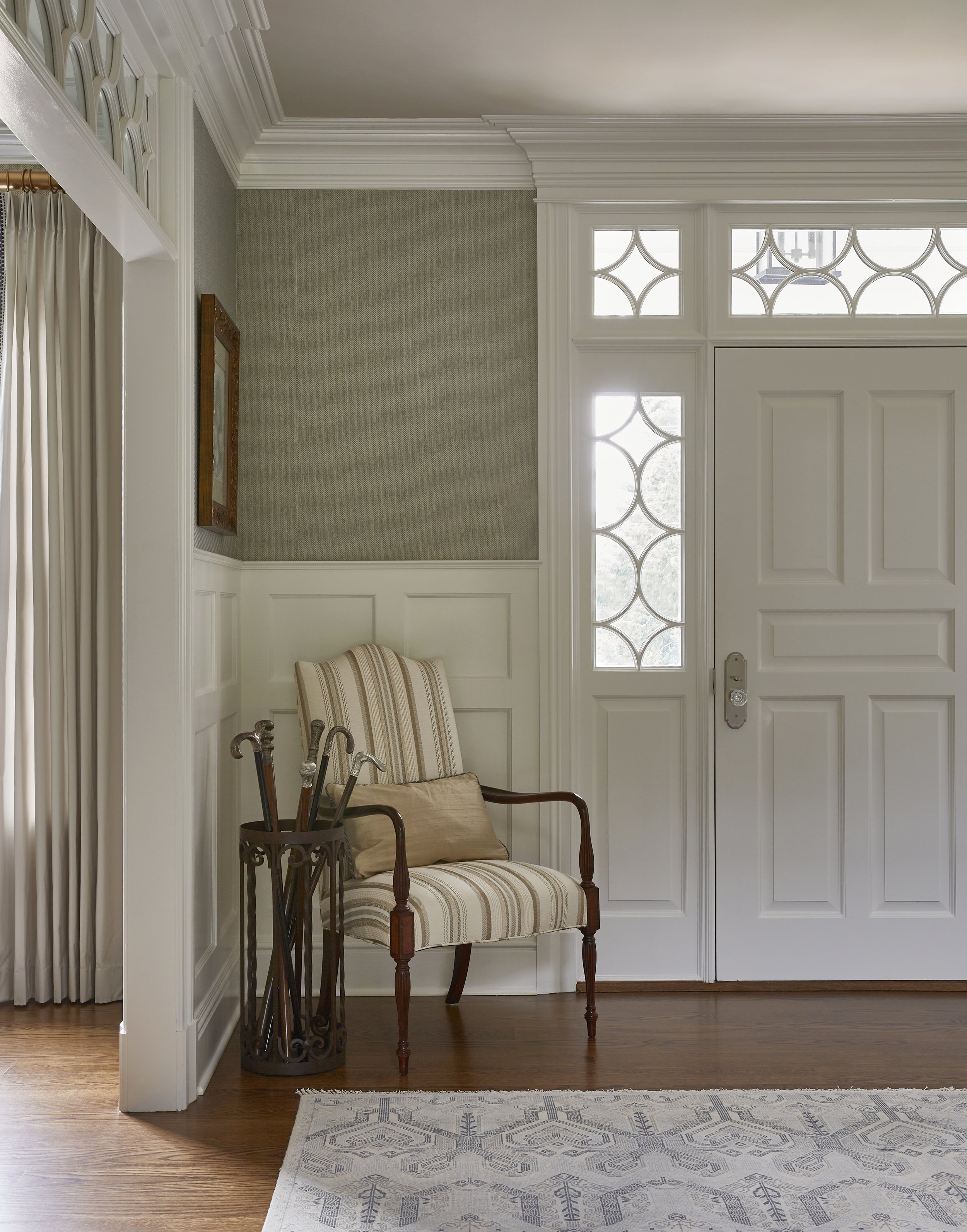 Entryway with a striped upholstered armchair, umbrella stand with umbrellas, a white door with decorative glass, hardwood flooring, and a patterned area rug