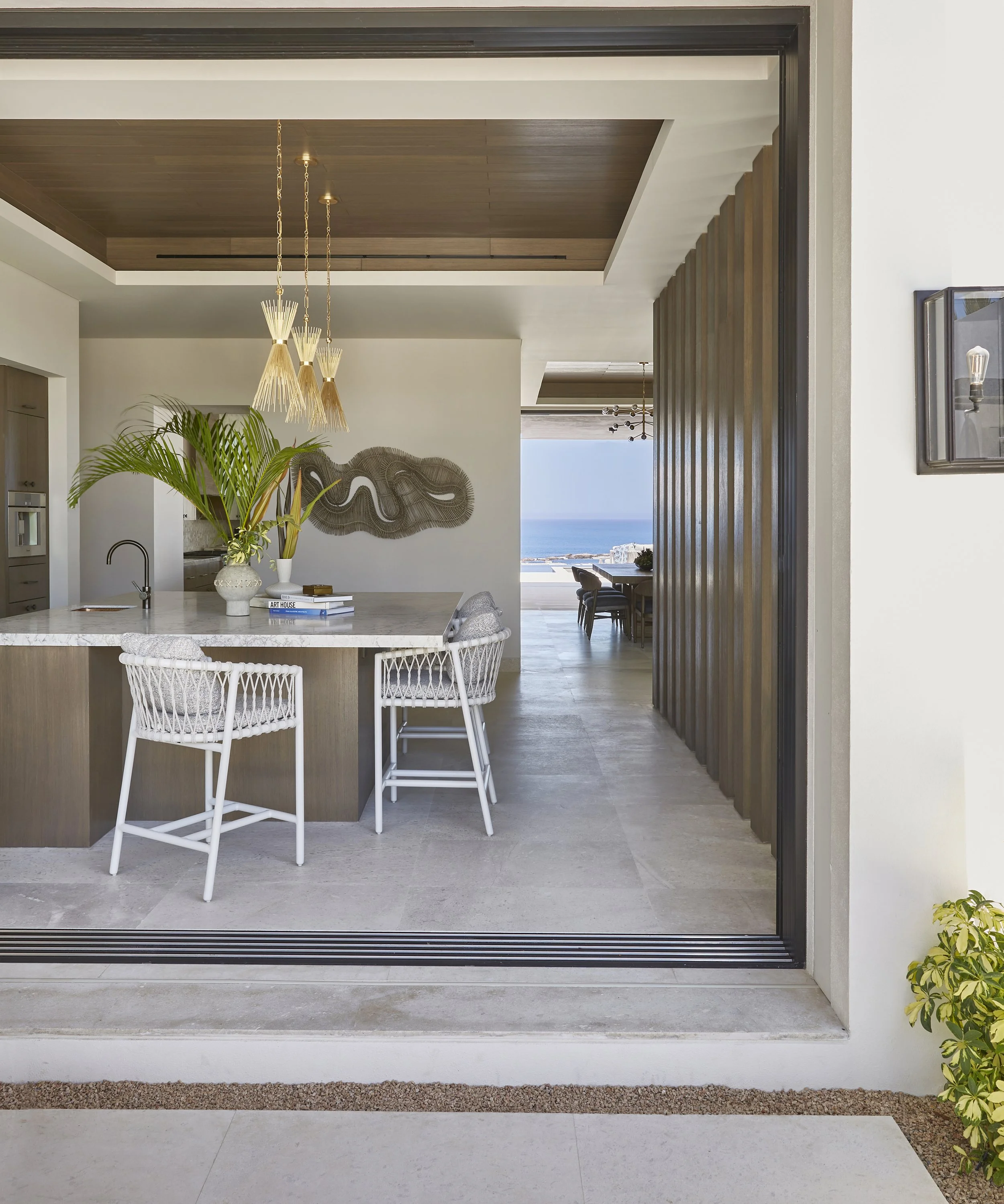 Modern open-concept kitchen with a marble island, white woven chairs, decorative wall art, green plants, and a view of the ocean in the background.