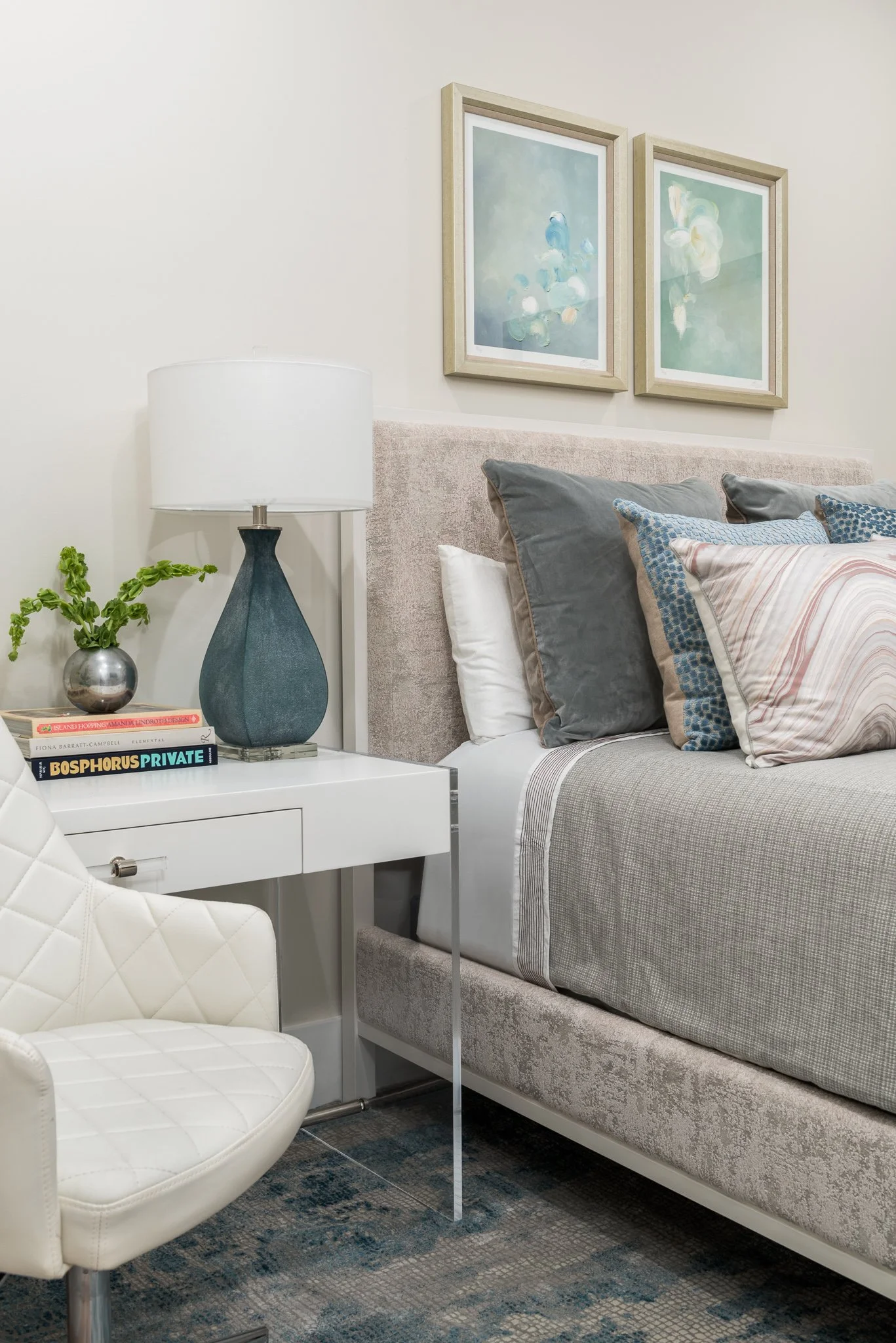 Close-up of a bedroom corner featuring a white nightstand with a blue table lamp, a small potted plant, and books. Part of a bed with a beige headboard, gray and patterned pillows, and framed abstract artwork hanging above.
