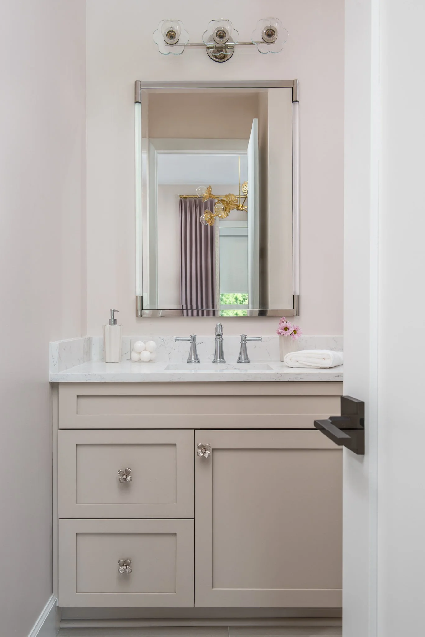 Bathroom vanity with a mirror, a sink, and decorative items including a soap dispenser, a container with balls, a flower vase, and a folded towel. A light fixture with three glass bulbs is above the mirror.