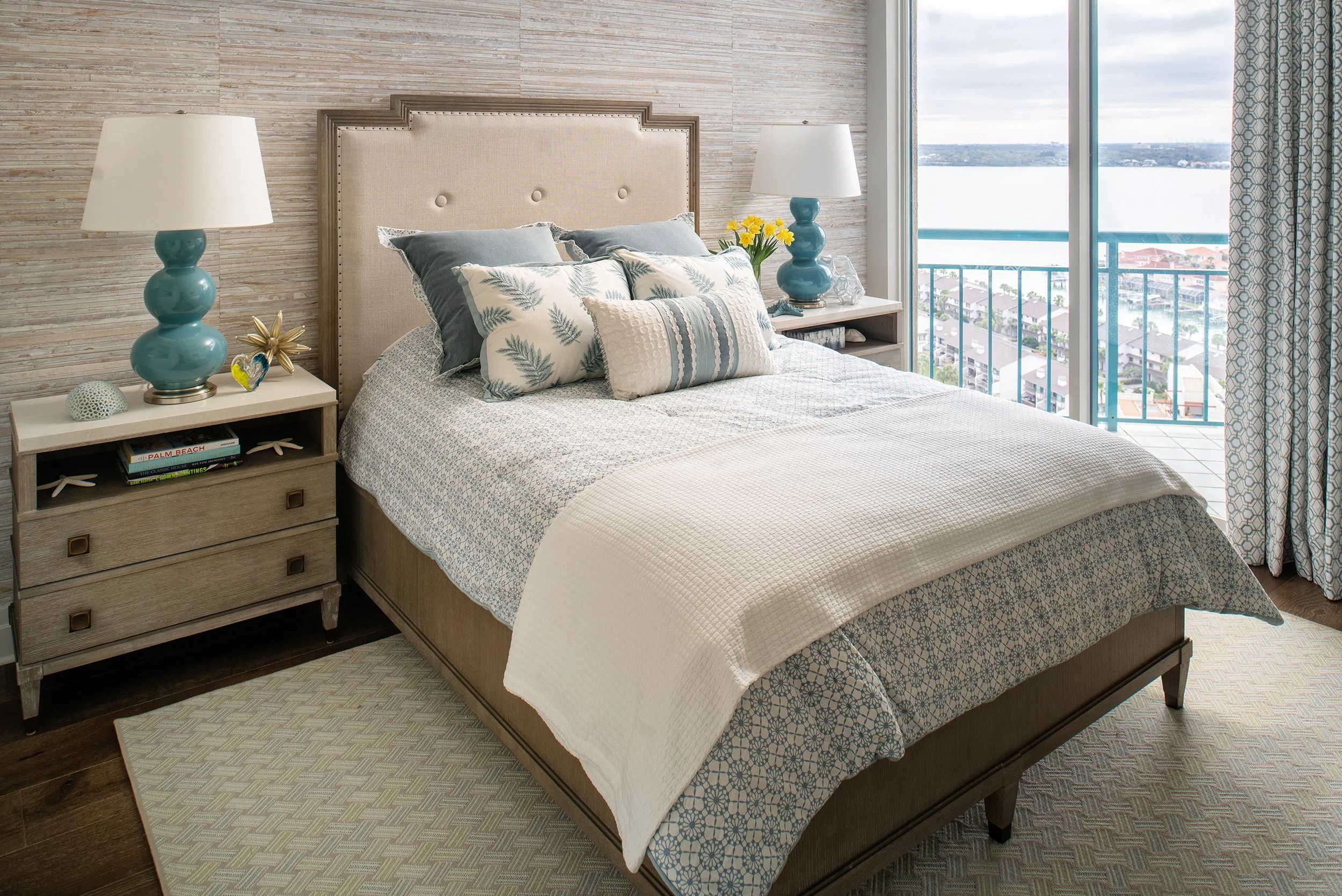 Beach-view bedroom with a bed, two nightstands with blue lamps, and large sliding glass doors opening to a balcony with a water and island view.