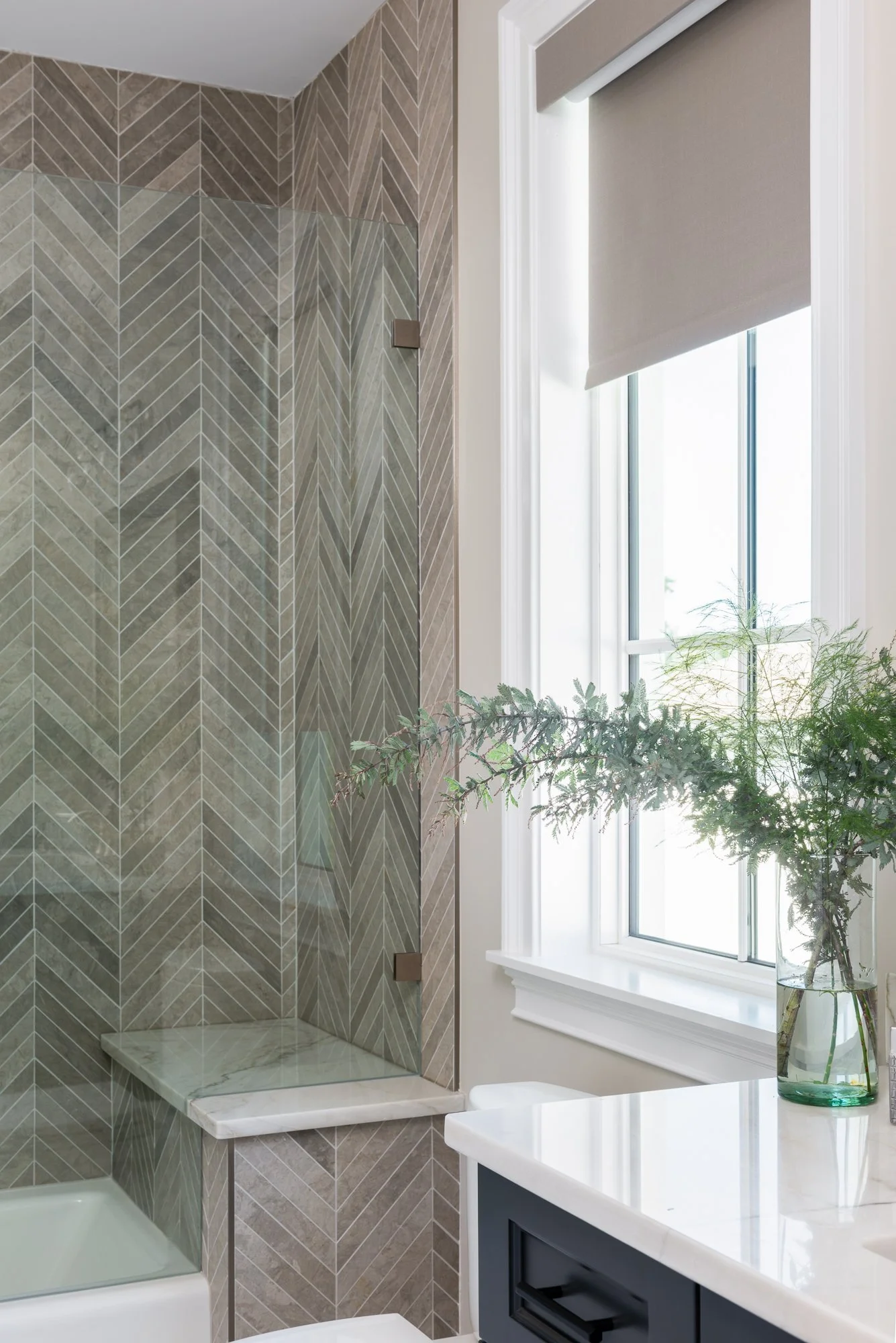 A bathroom with a khaki-colored chevron-tiled shower, a white countertop, a navy blue cabinet, and a window with a beige shade, decorated with a glass vase and green foliage.