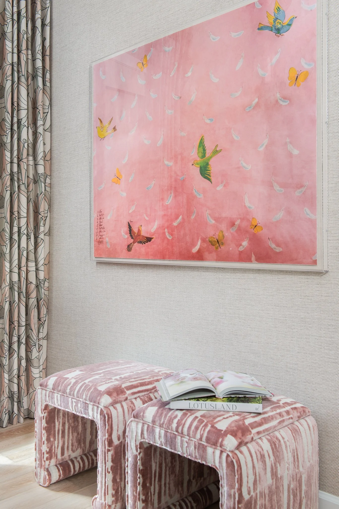 A pink abstract painting with colorful birds and butterflies hangs on a wall in a room with patterned curtains. Two pink velvet stools with an open magazine on top are in the foreground.