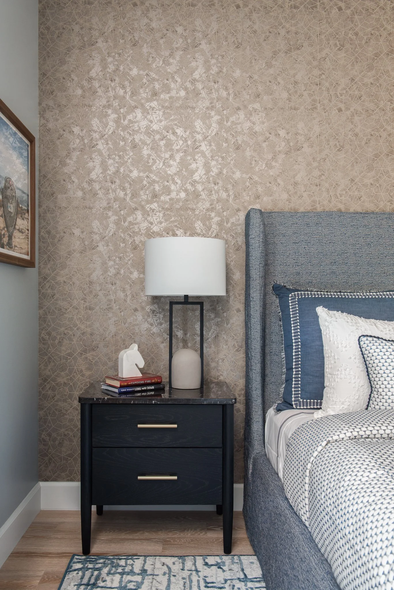 Bedroom nightstand with a white lamp, a white decorative horse, and a stack of books next to a bed with blue and white bedding, against a textured beige wall.