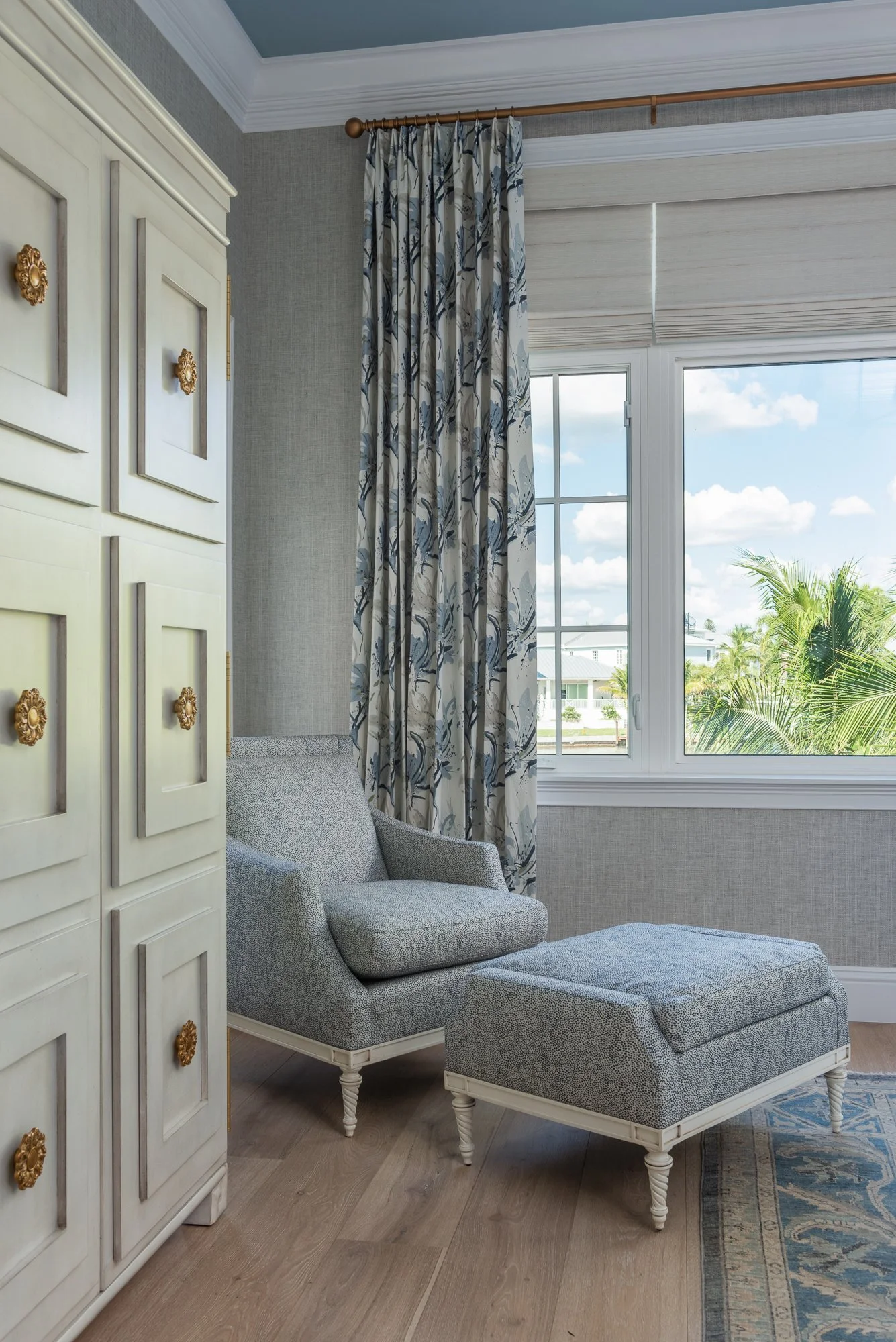 A corner of a room with a white wardrobe, a gray armchair with an ottoman, large window with floral curtains, and a view of palm trees outside.