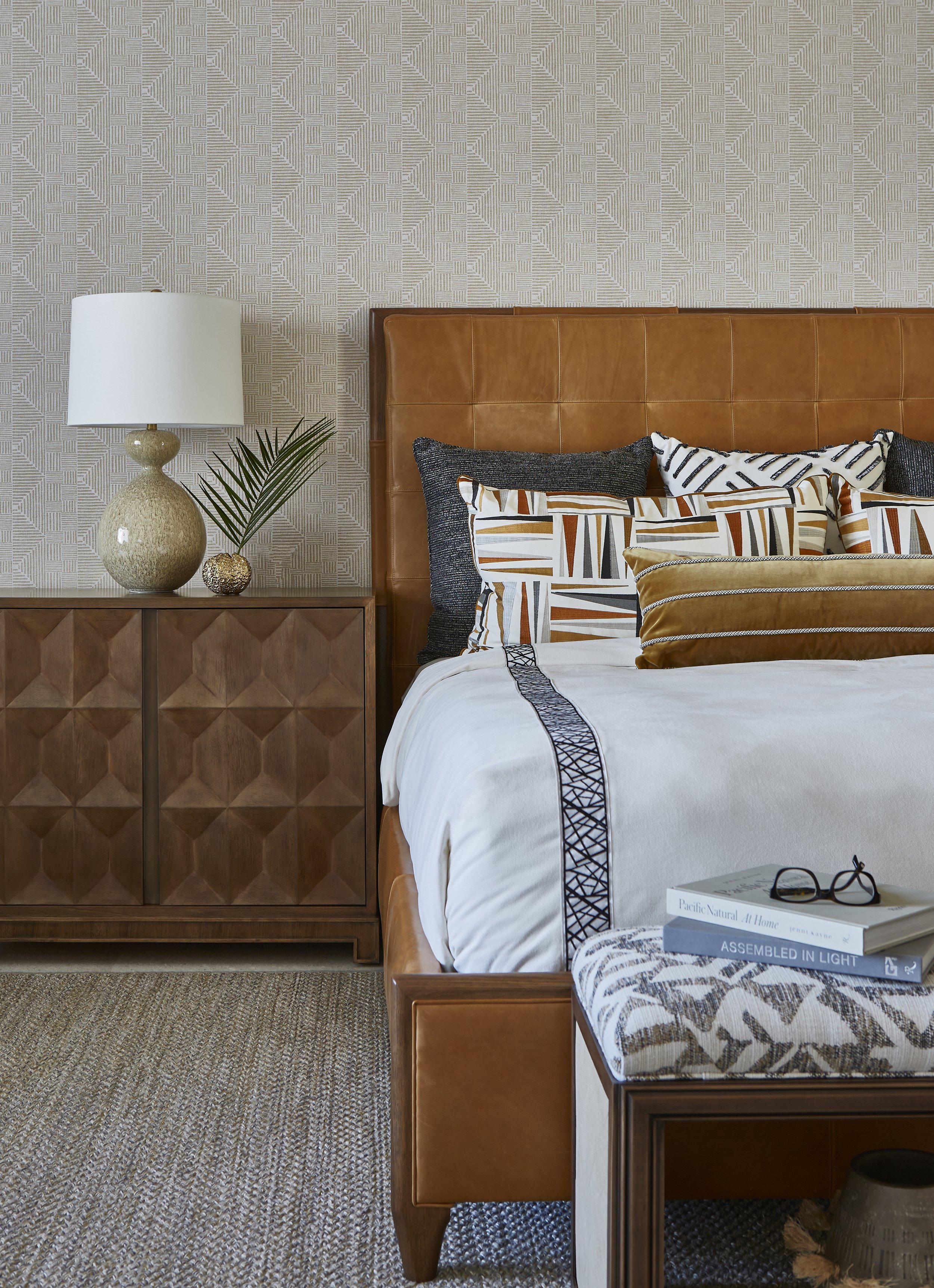 A bedroom with a tan leather headboard, patterned pillows, a wooden nightstand with a lamp, a decorative branch, and books on a matching bench.