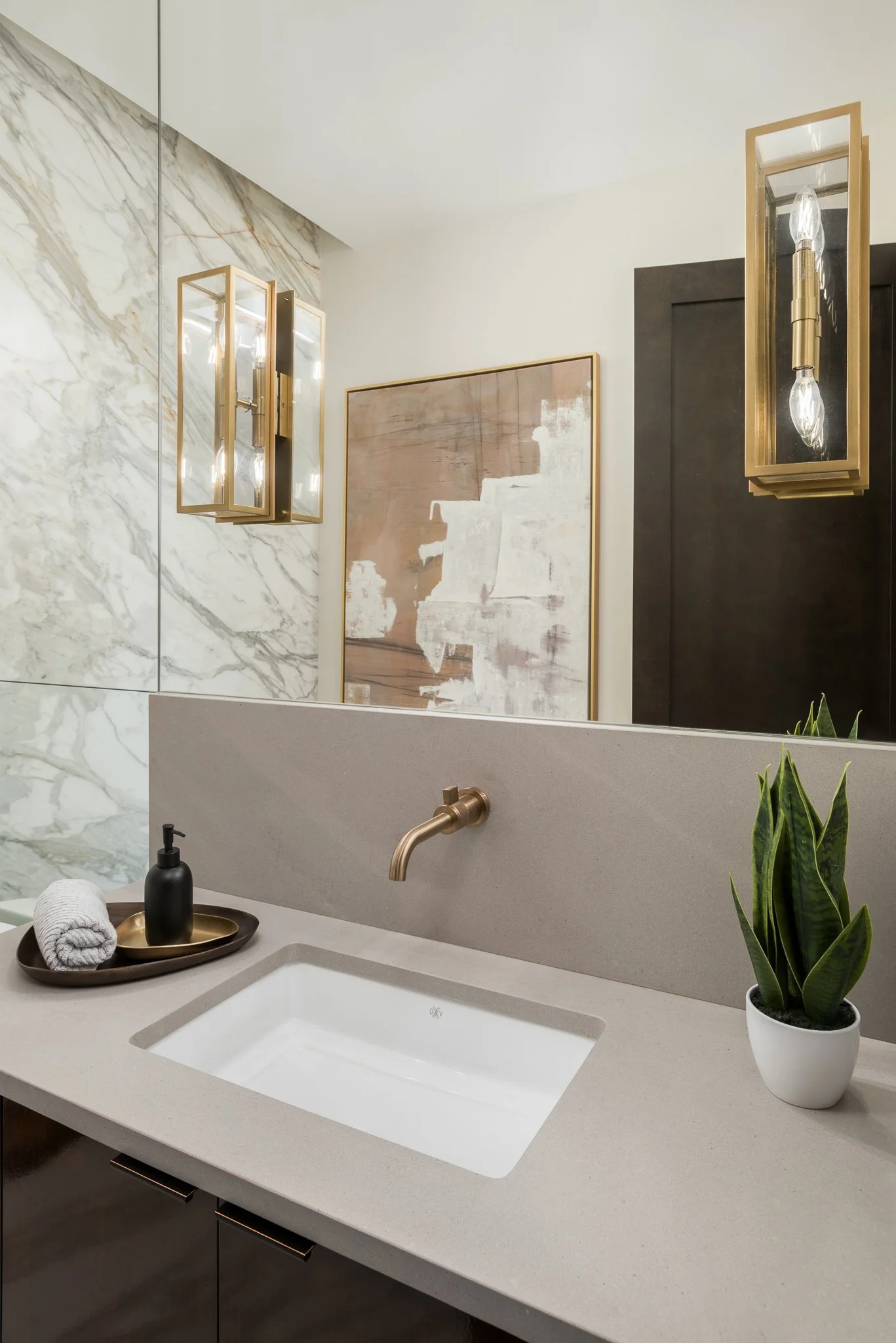 A modern bathroom with a beige countertop, a white rectangular sink, a gold faucet, a black soap dispenser, a white towel, and a gold tray. There is a potted green plant on the right side of the counter. The wall behind the sink has a large mirror an