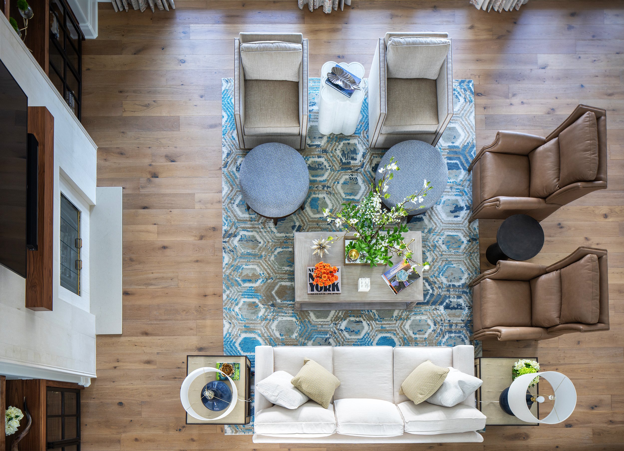 Top-down view of a living room with a white sofa, a coffee table with flowers and magazines, two beige armchairs, two gray ottomans, a brown recliner, a black side table, a fireplace, and a rug with a hexagonal pattern.
