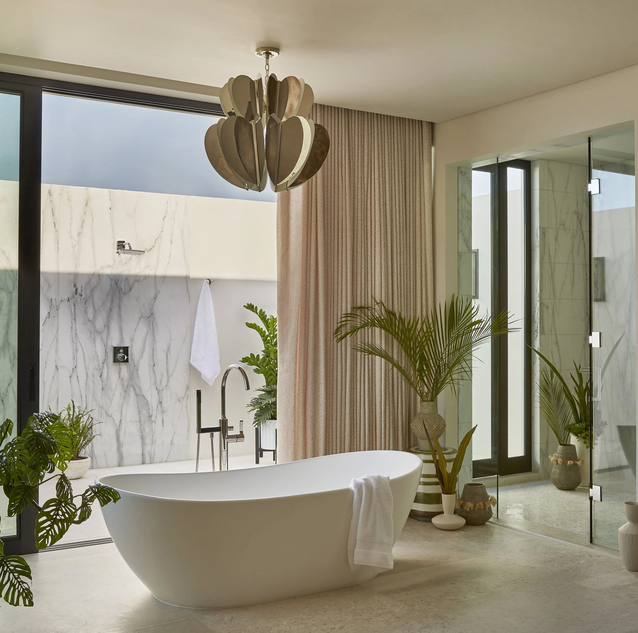 Modern bathroom with a freestanding bathtub, large window, marble accents, potted plants, and a decorative ceiling light.