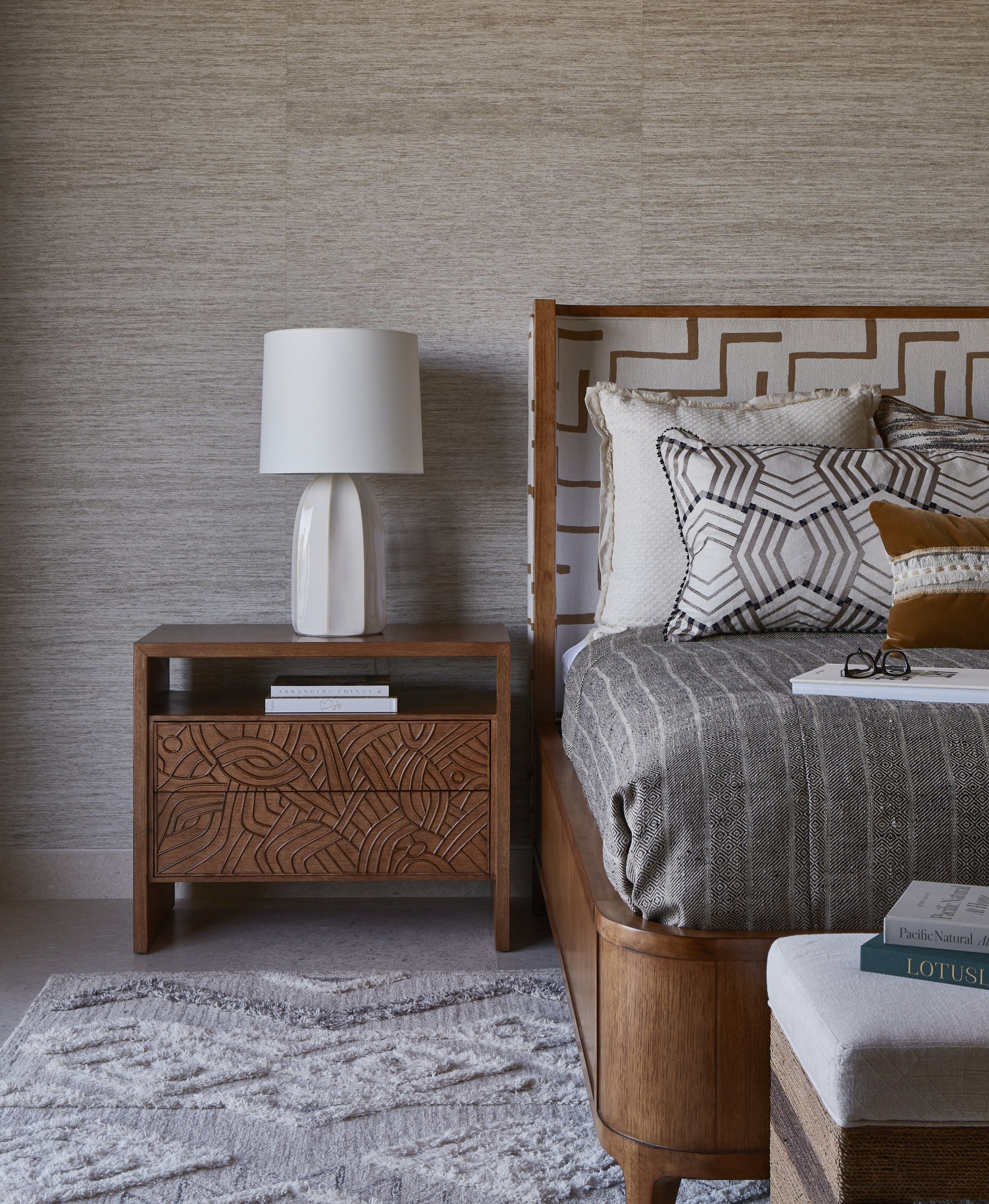 A bedroom with a wooden nightstand, a white lamp, and several pillows on a bed with patterned bedding. There are books on both the nightstand and a nearby bench or ottoman.