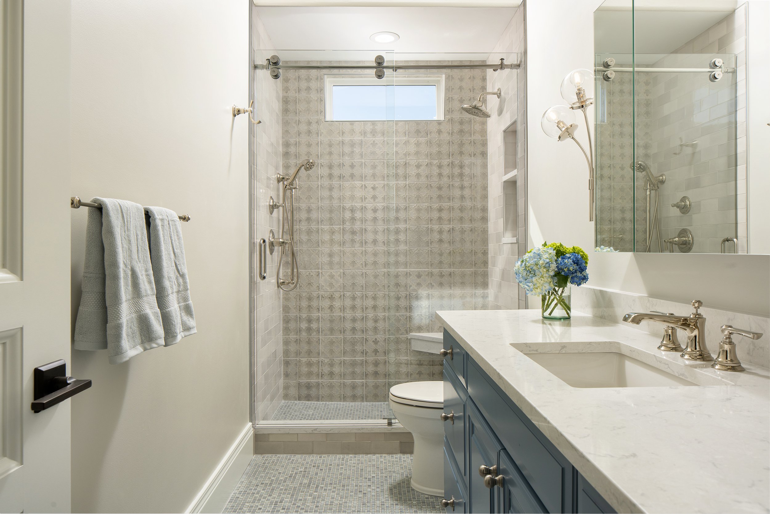 A modern bathroom with a glass-enclosed shower, a white marble countertop with a sink, and blue cabinetry. There is a vase of blue and white hydrangeas on the counter, and towels hanging on a silver towel bar. The shower has a window at the top and d
