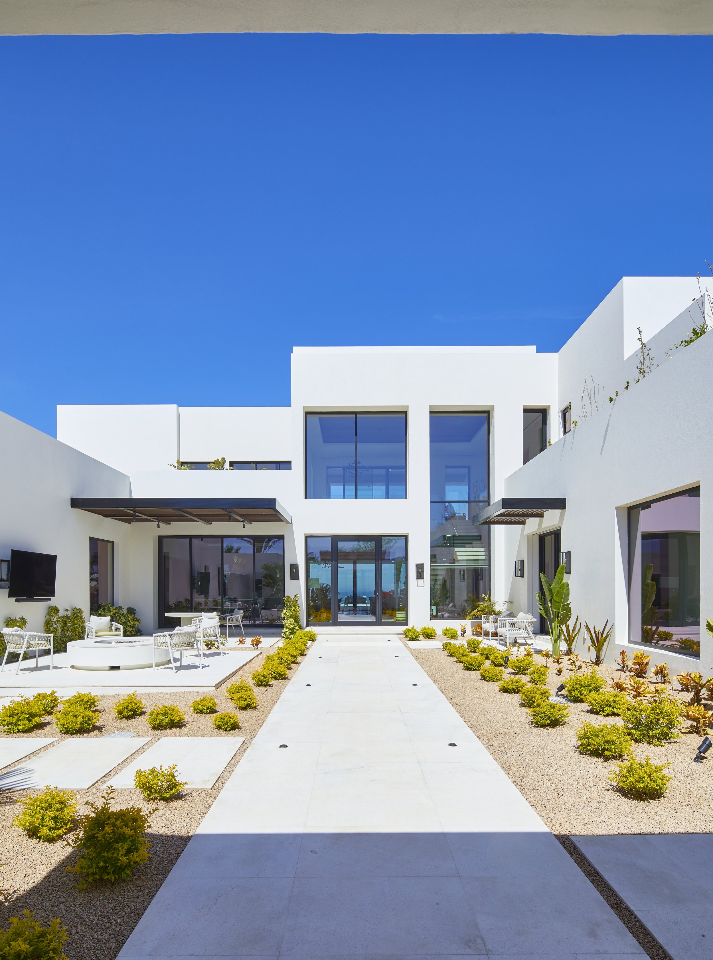 Modern white house with large windows and outdoor seating area, landscaped with small bushes and gravel, against a bright blue sky.