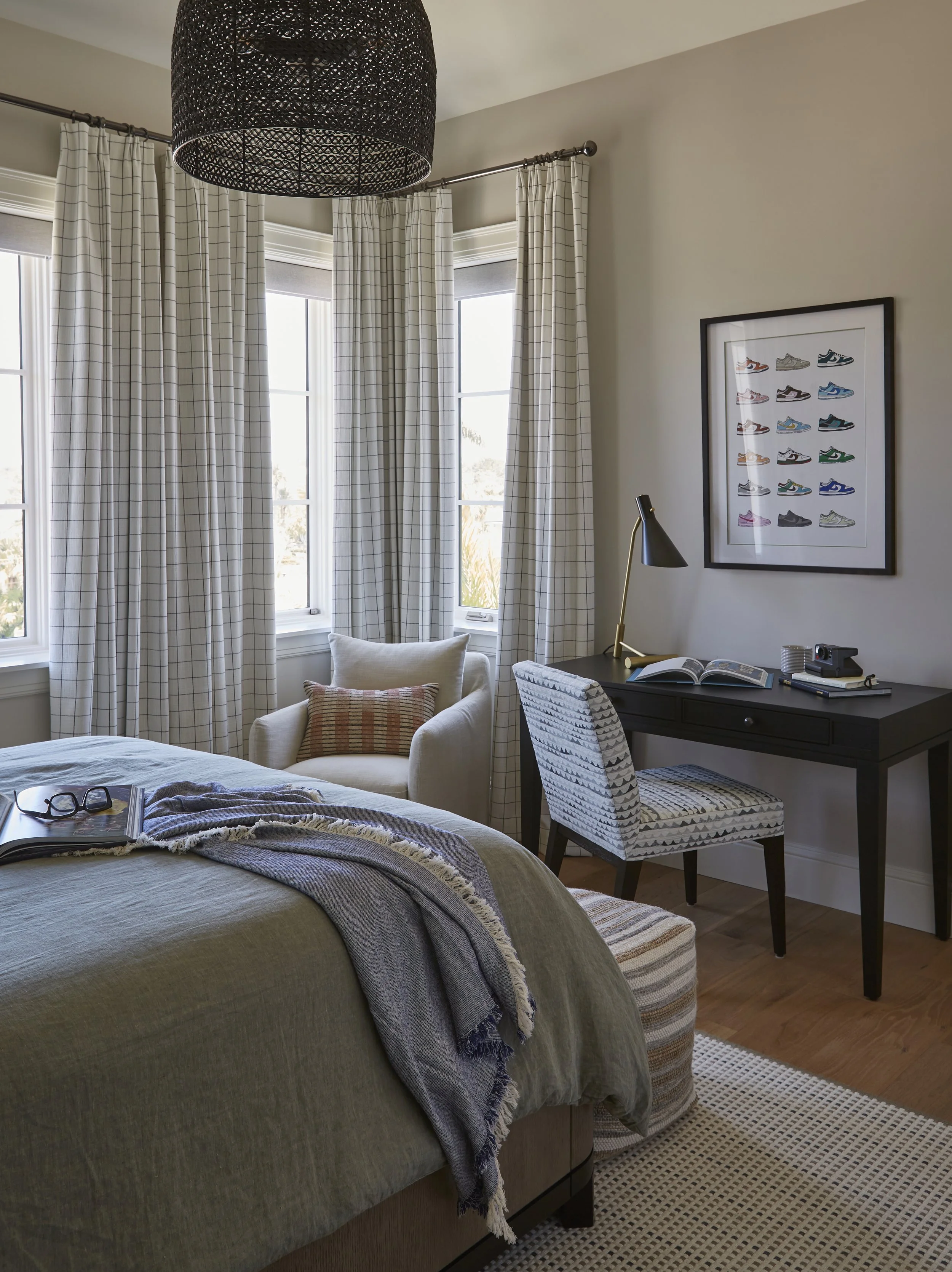A cozy bedroom with a bed, a white armchair, a black desk, and a framed collection of colorful sneakers on the wall. The room is bright with three windows and white checkered curtains.