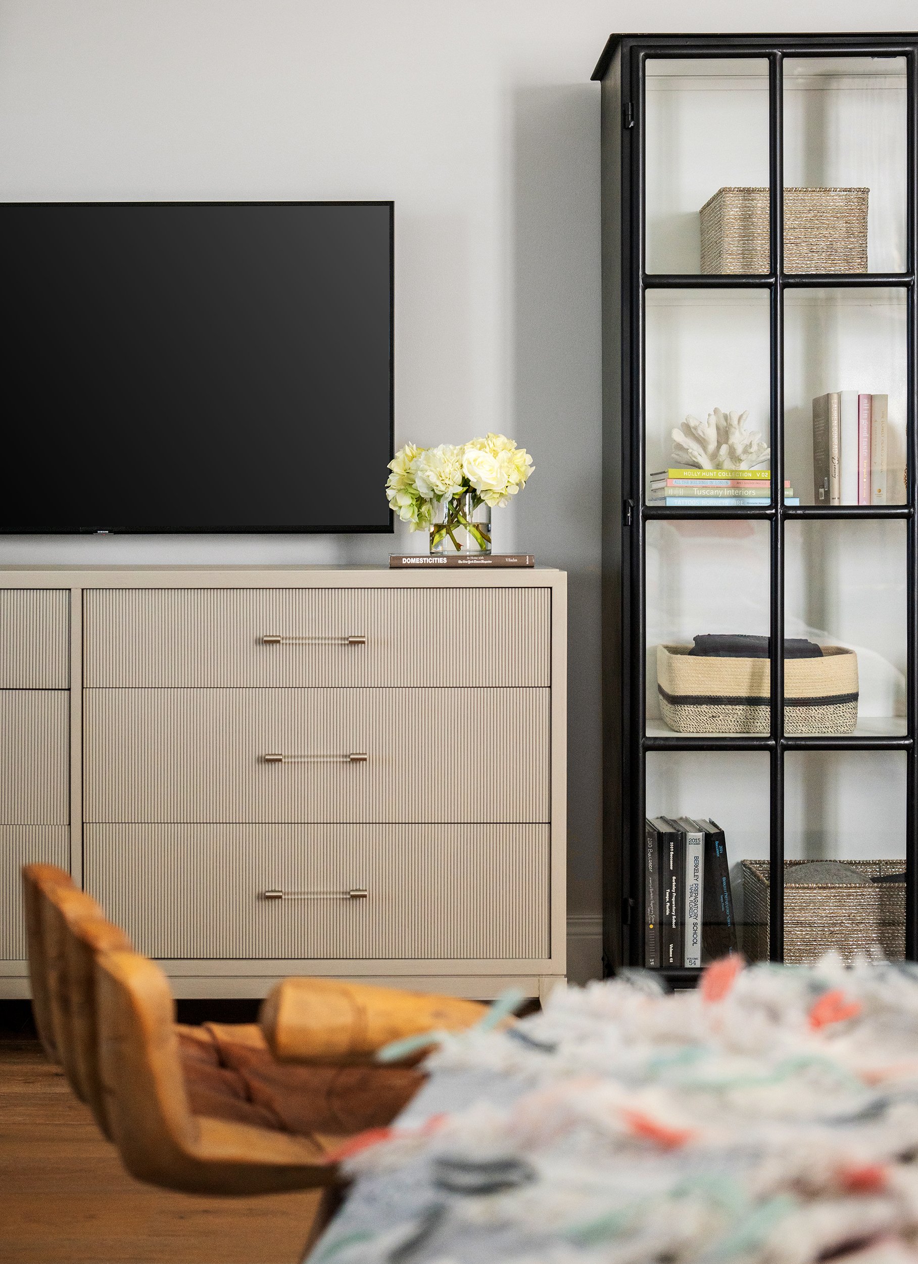 Living room with a beige dresser holding a vase of white flowers, a black frame glass cabinet containing books and decorative items, and a blurred wooden chair in the foreground.
