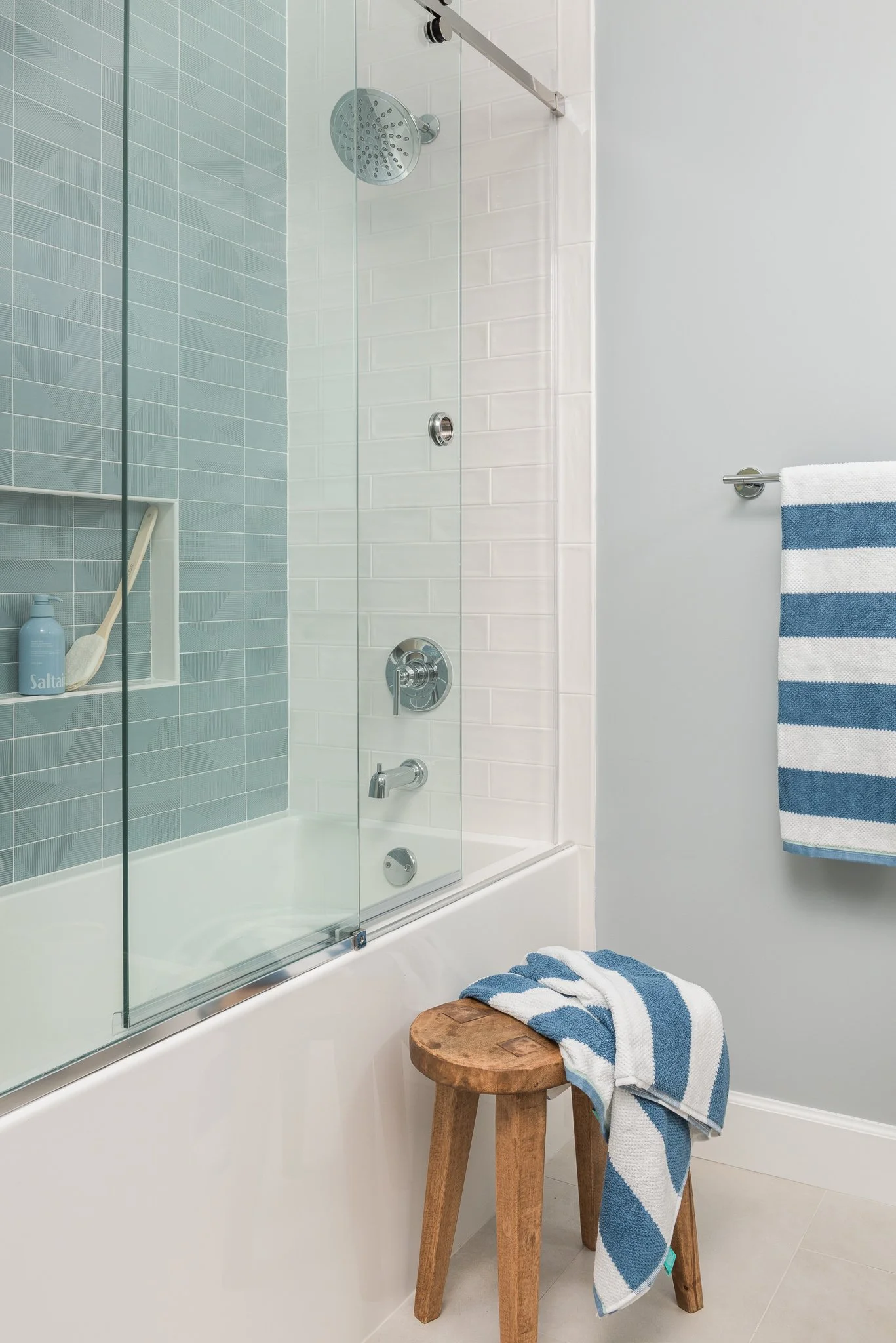 Bathroom with a bathtub, glass shower door, gray tiled wall, and a towel hanging on a rack. There is a small wooden stool with a blue and white striped towel draped over it.