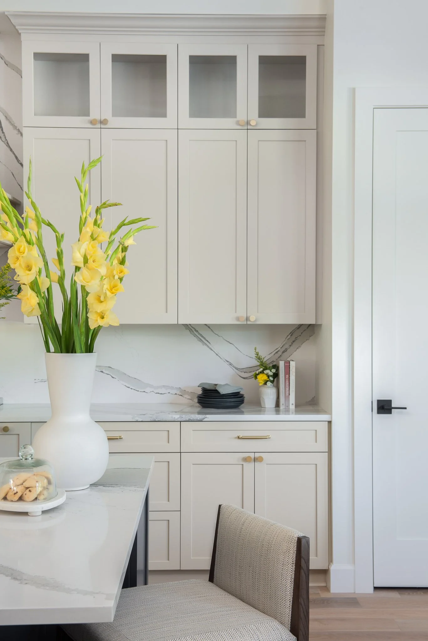 Interior view of a kitchen with white cabinets, a marble backsplash, a white table with a large yellow flower arrangement in a white vase, and a beige upholstered chair.