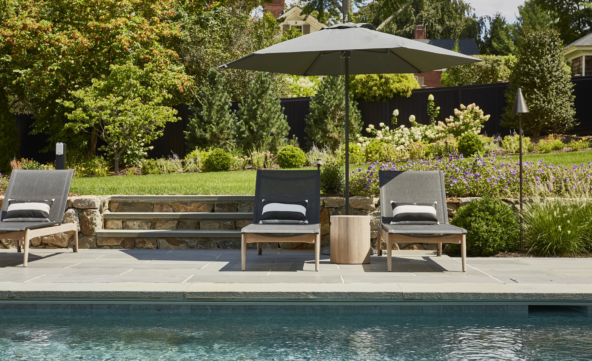 Backyard with pool, patio, garden, three lounge chairs with small pillows, large umbrella, small side table, and flowering bushes and trees in the background.