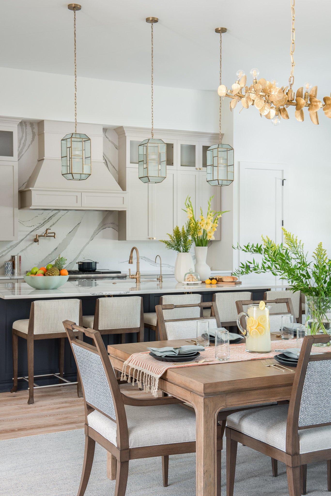 Modern kitchen and dining area with pendant lights, a wooden dining table with chairs, and a kitchen island with chairs, decorated with green plants and flowers.