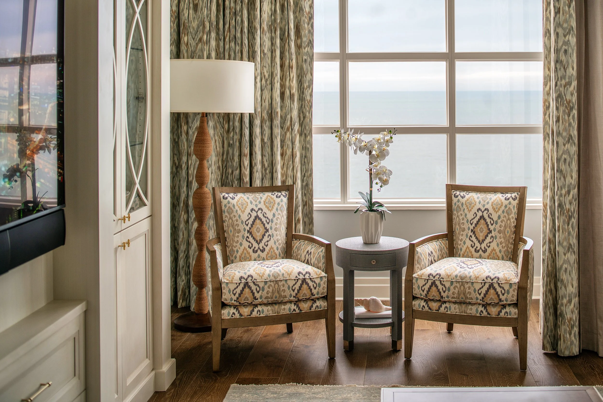 A cozy sitting area with two patterned armchairs, a small round table with a vase of white orchids, and a large window showing an ocean view.
