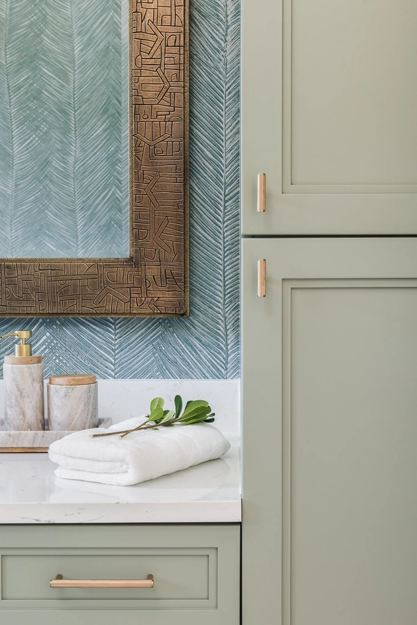 Close-up of a bathroom vanity with sage green cabinets, a white marble countertop, a rolled white towel with a small branch, a soap dispenser, and a matching tissue holder. A framed mirror with a wooden carved border hangs on a blue and green pattern