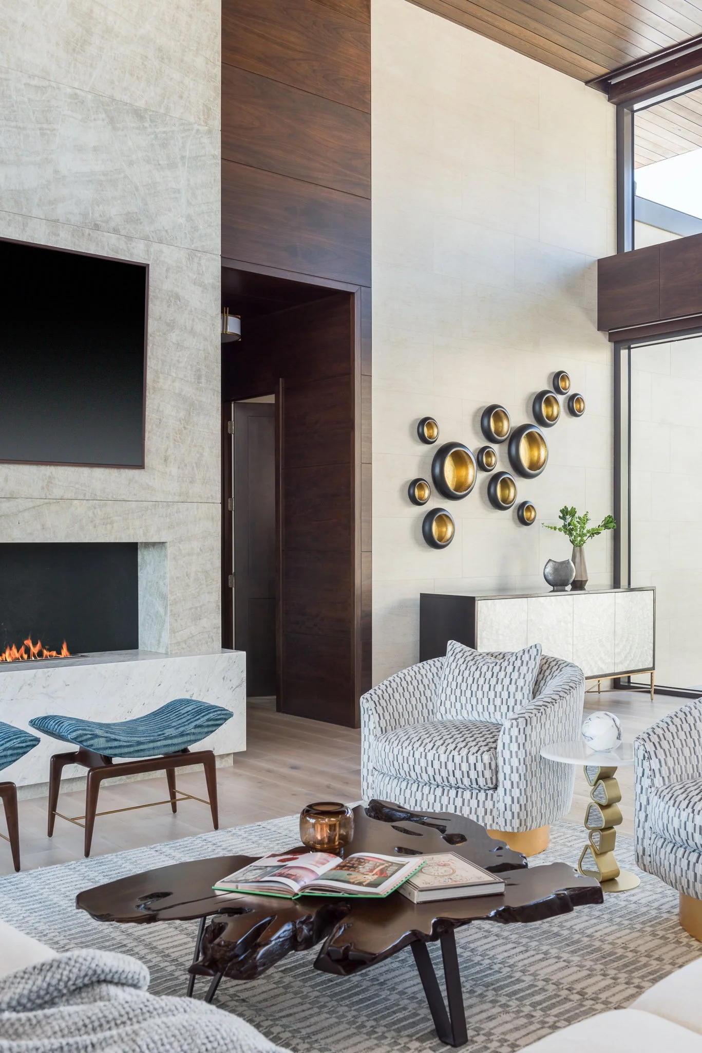 Modern living room with a fireplace, wall-mounted TV, decorative wall art, patterned chairs, coffee table with books, and a side table with a sculpture, featuring large windows and wooden ceiling.