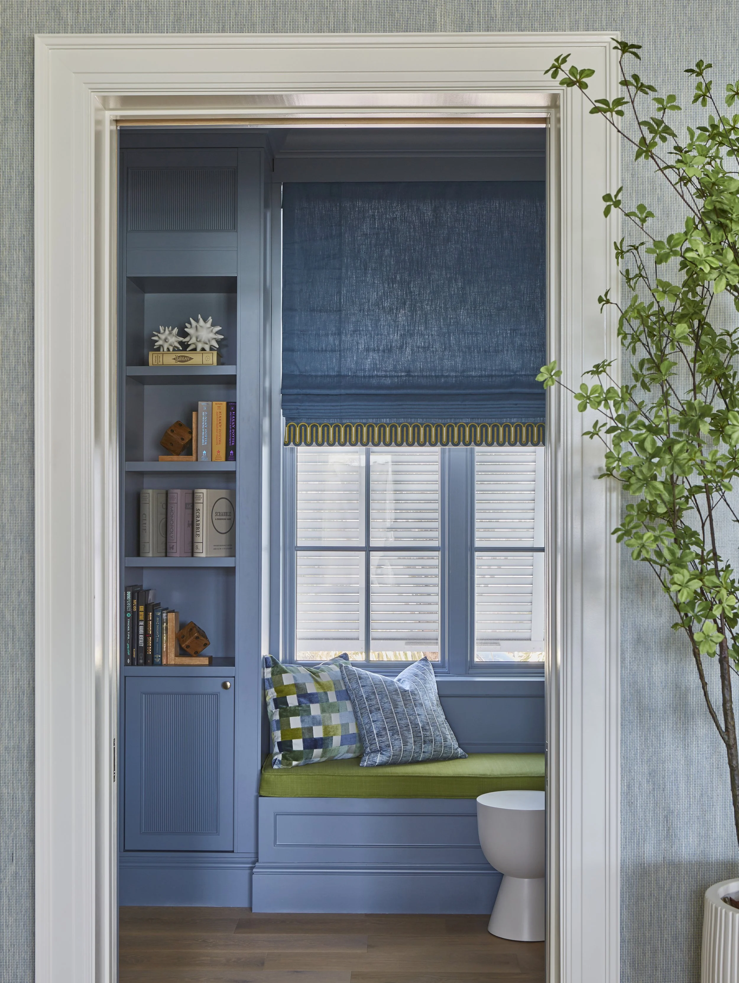 A cozy window nook with a blue built-in bench, two plaid pillows, a large window with closed blinds, a blue roller shade, bookshelf with books and decorative objects, and a potted plant nearby.