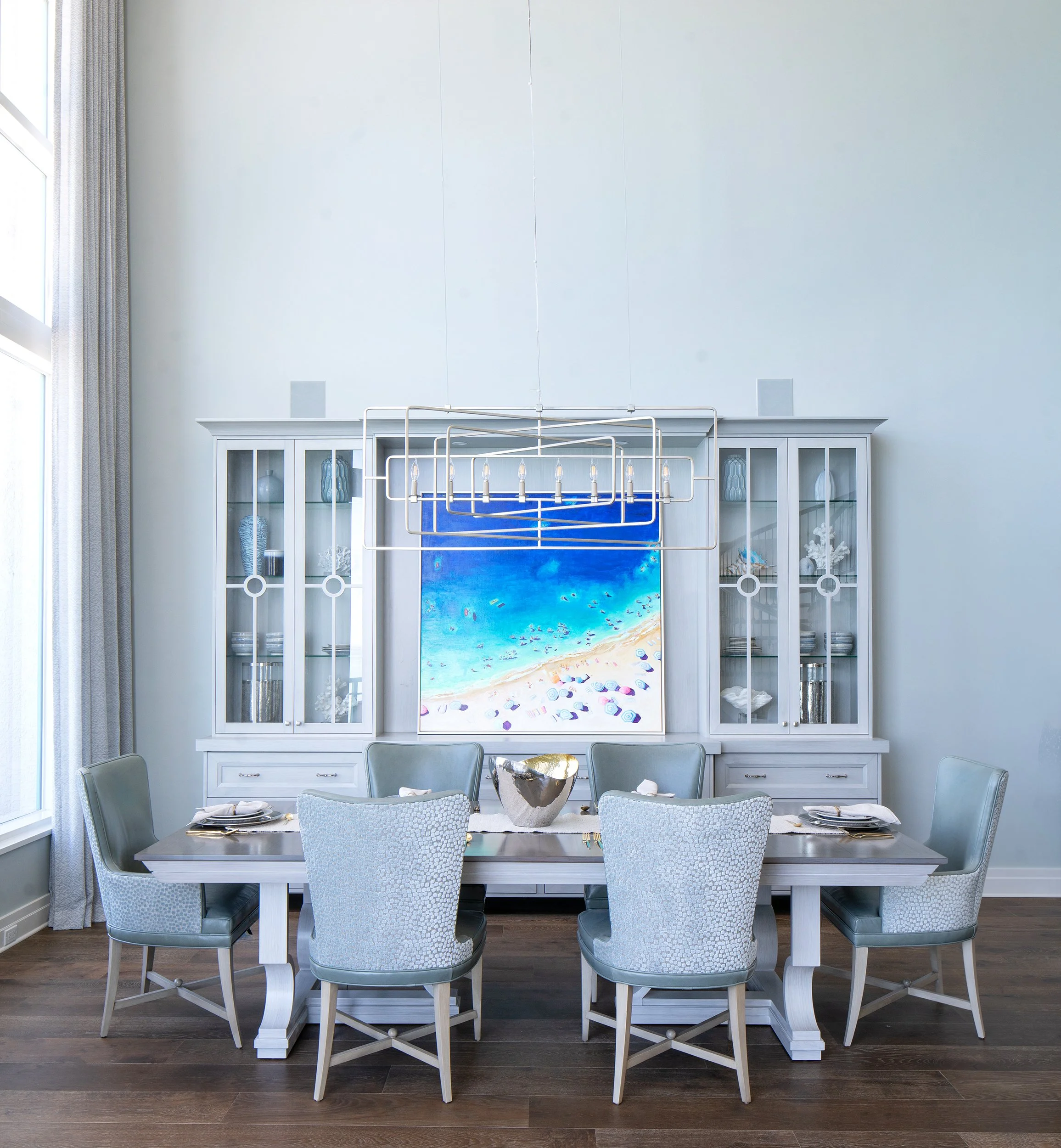 A dining room with a large white table, six upholstered chairs, a white china cabinet displaying dishes and vases, a beach-themed painting, and a modern geometric chandelier.