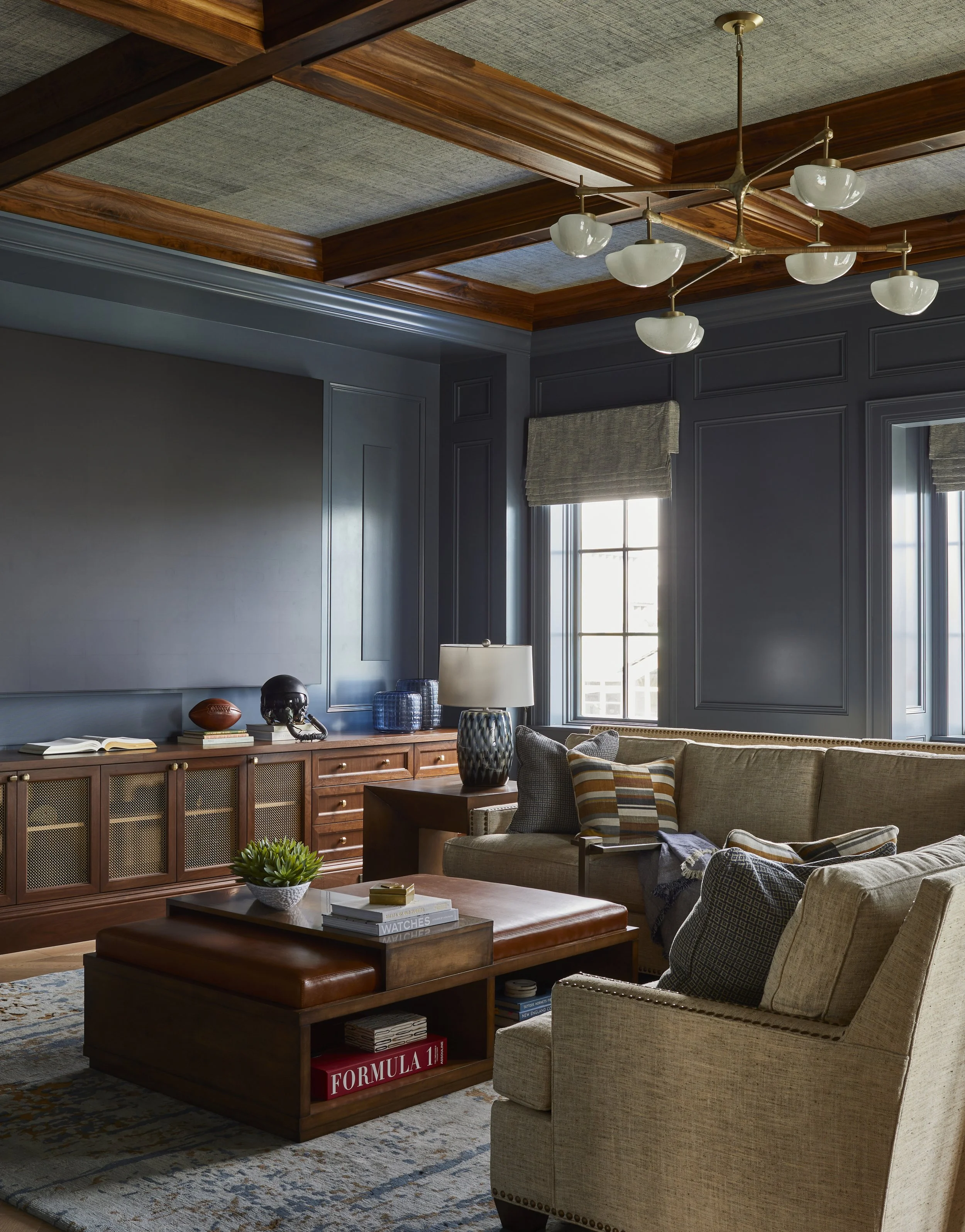 Living room with dark blue walls, wooden ceiling, beige sofas, wooden coffee table, area rug, and windows with gray shades.