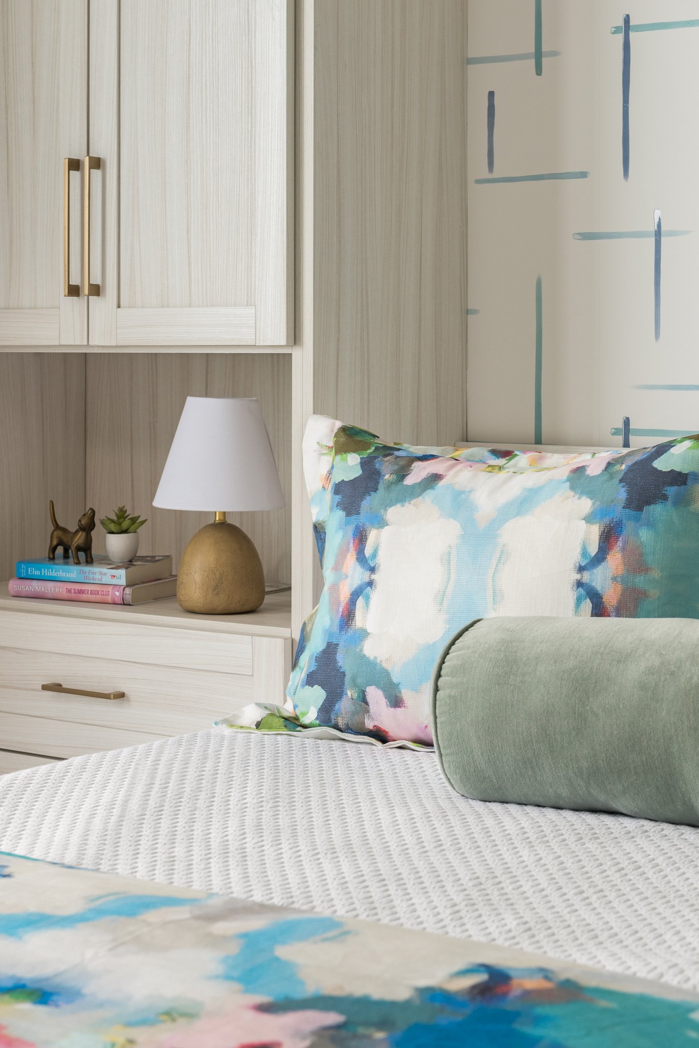 Close-up of a bed with a multifaceted decorative pillow, a soft beige blanket, and a colorful floral bedspread in a bedroom with light wood cabinetry and a nightstand with a white lamp, books, and decorative items.