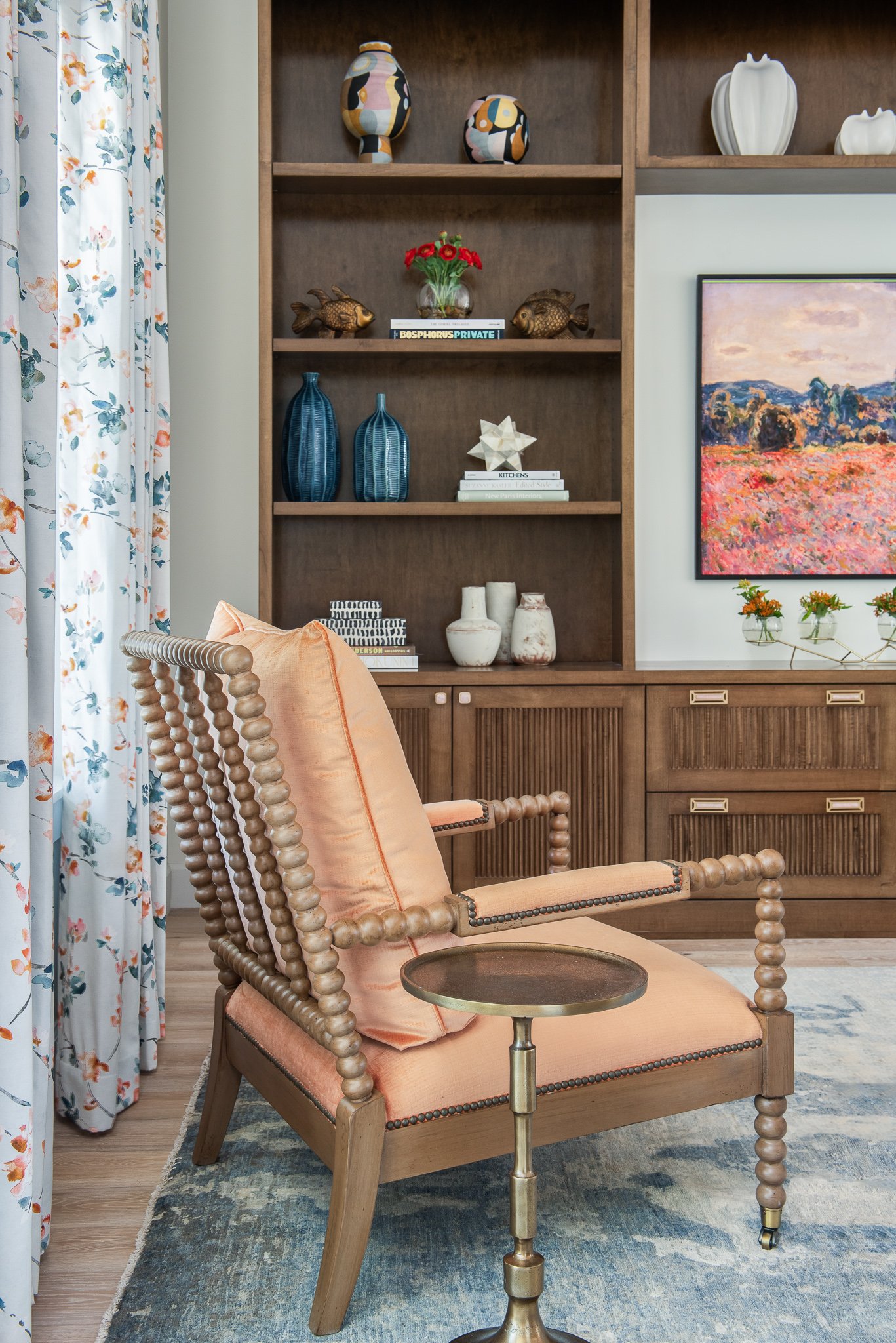 Living room with a beige armchair, a side table, a wooden bookshelf with decorative items, a colorful floral curtain, a colorful landscape painting, and small vases with flowers.