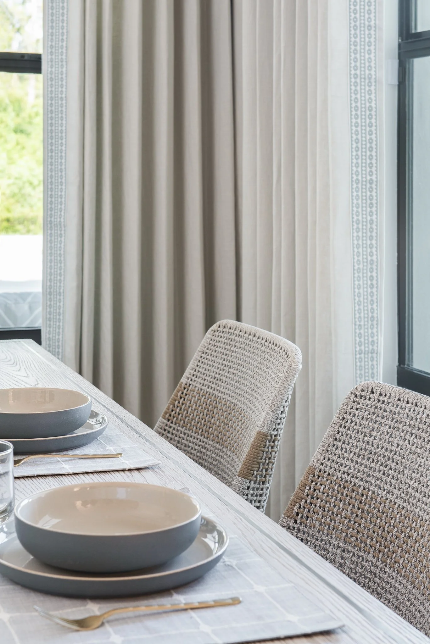 Set dining table with bowls, plates, and utensils in a bright room with beige curtains and woven chairs.