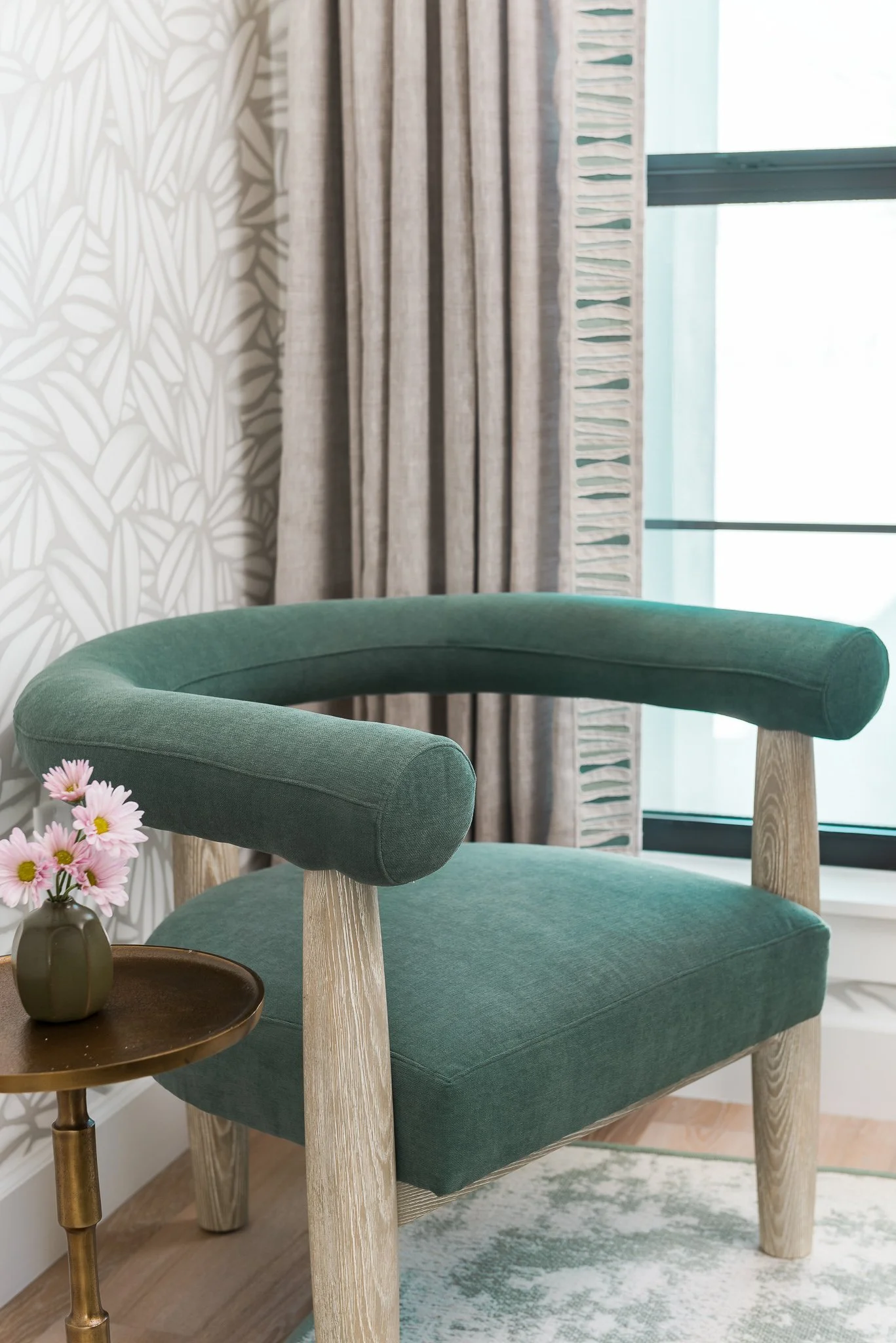 Green upholstered armchair with wooden legs in a room with patterned wallpaper, beige curtains, and a window. A small side table with pink flowers in a vase is nearby.