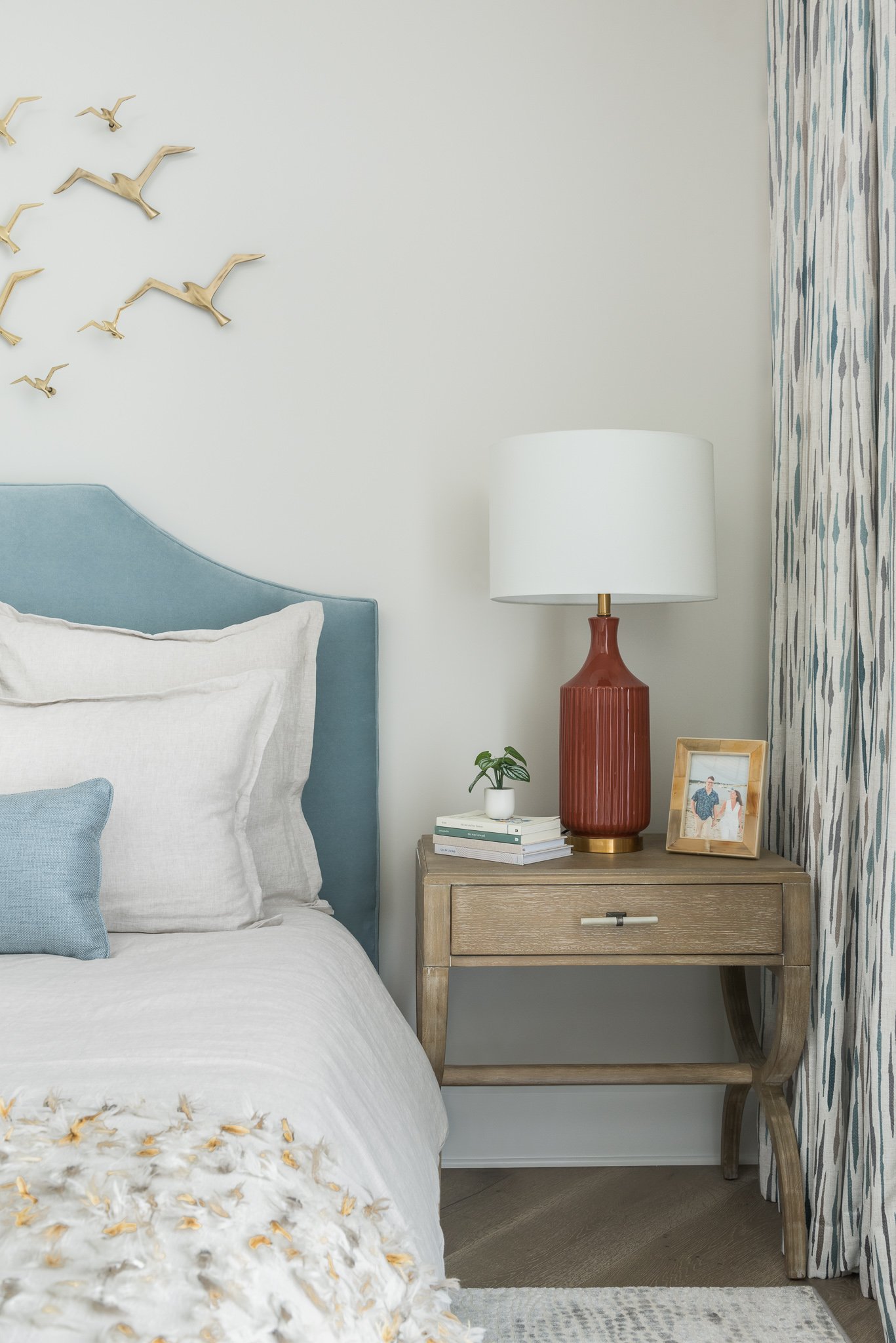A bedroom scene with a bed, pillows, a nightstand, a table lamp, a small plant, a framed photo, and decorative bird wall art.