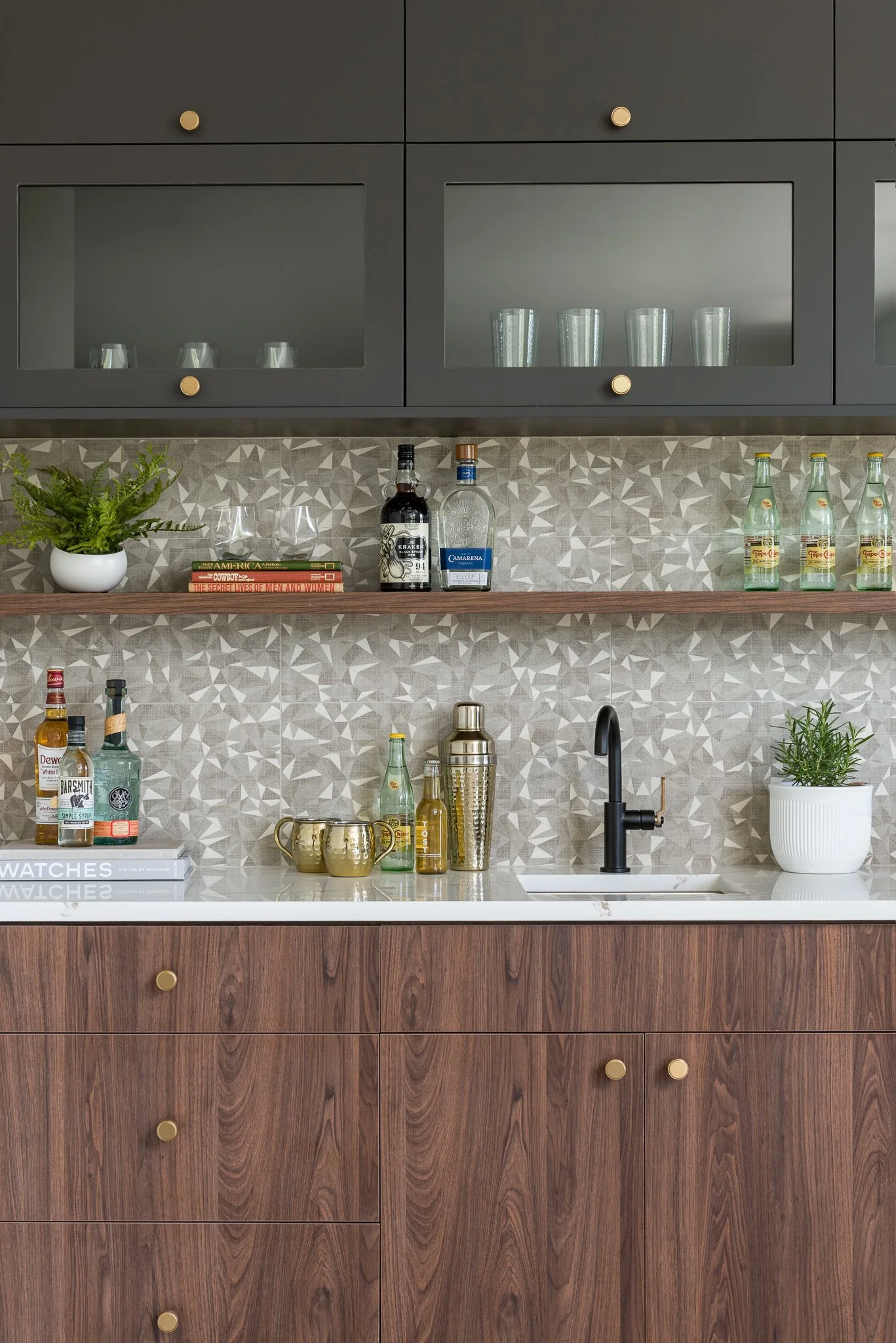 Modern kitchen with dark upper cabinets, wooden lower cabinets, a patterned backsplash, a black faucet, and various bottles, glasses, and plants on the countertop and shelves.