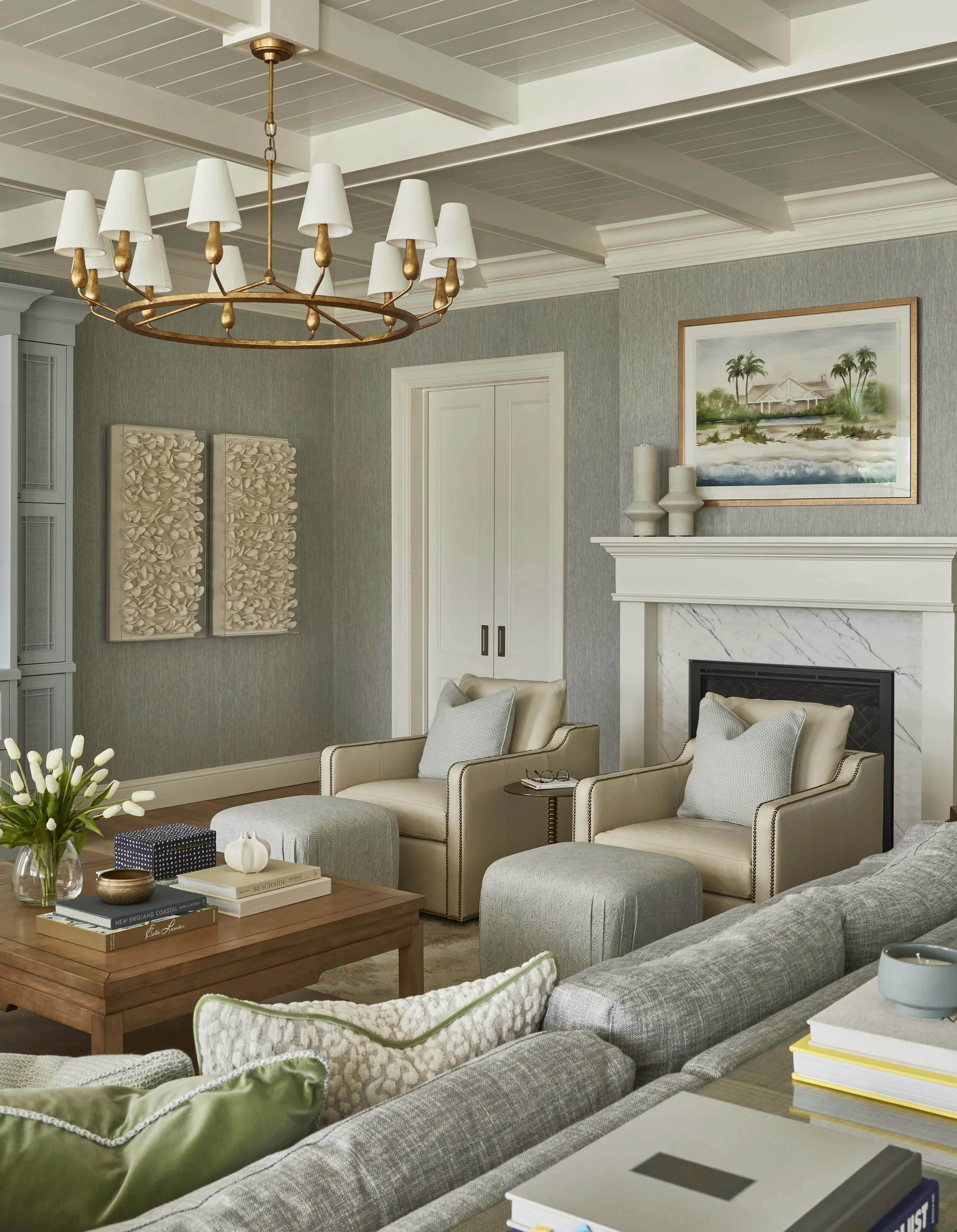 A living room with a chandelier, framed artwork, fireplace, armchairs with pillows, a sofa with pillows, a coffee table with decorative items, and a fireplace.