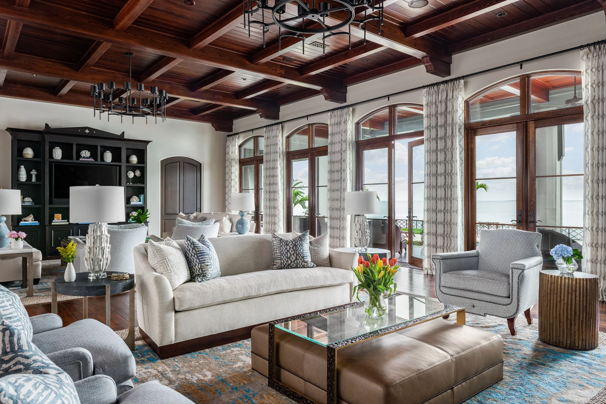 A spacious living room with large arched windows and wooden ceiling beams. Features cream-colored sofas, patterned armchairs, a glass-topped coffee table with a flower arrangement, and built-in shelves. The room opens to a balcony overlooking the oce