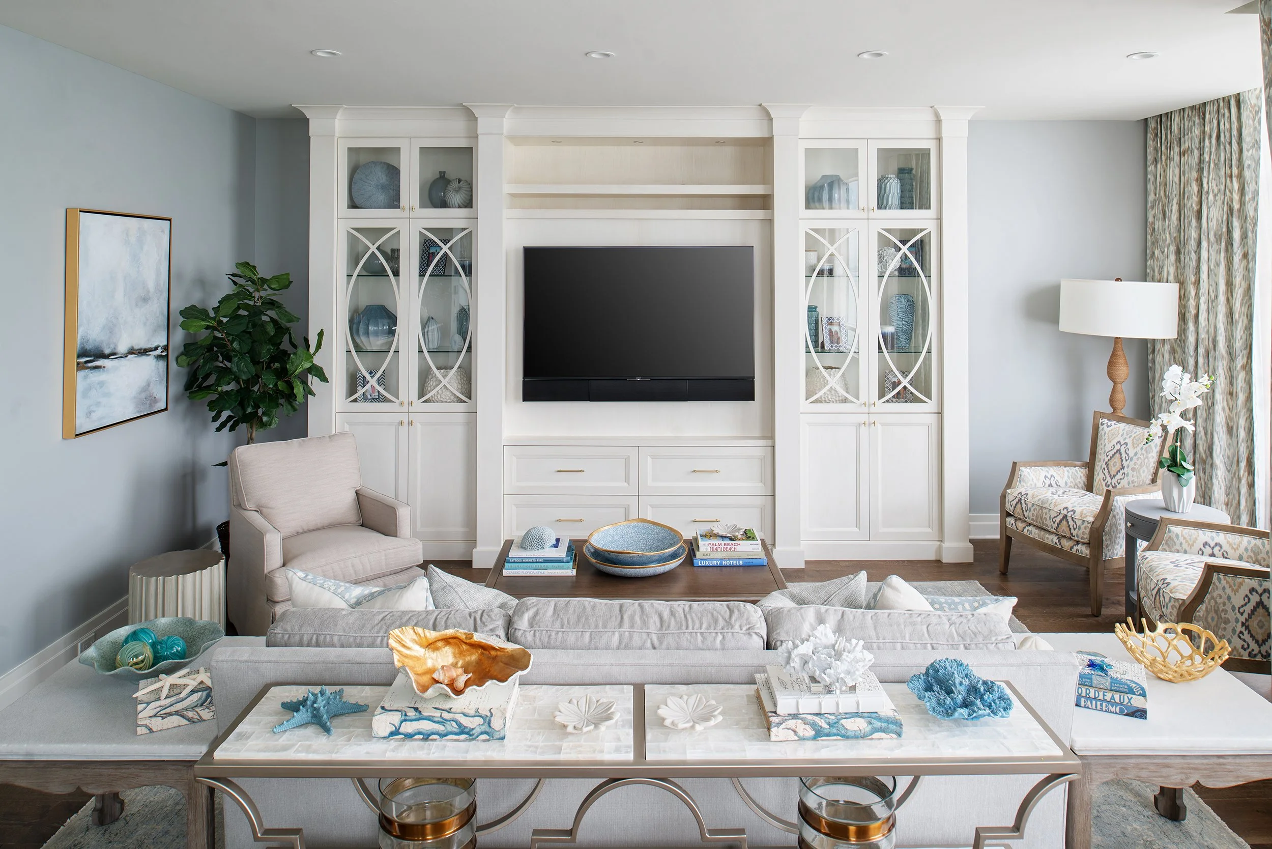 Living room with white built-in shelves, a flat-screen TV, beige armchair, patterned chairs, coffee table with decorations, and large window with floral curtains.
