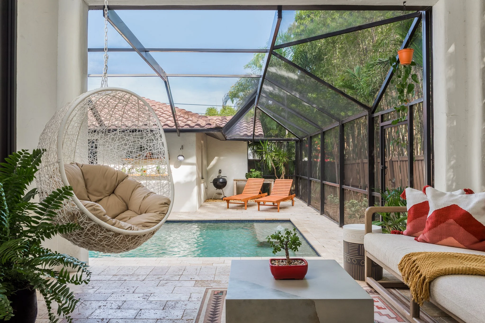 Indoor-outdoor patio with pool, wicker hanging chair, wooden lounge chairs, sofa with red pillows, potted plants, and a screened enclosure, overlooking a garden with trees and a tiled roof