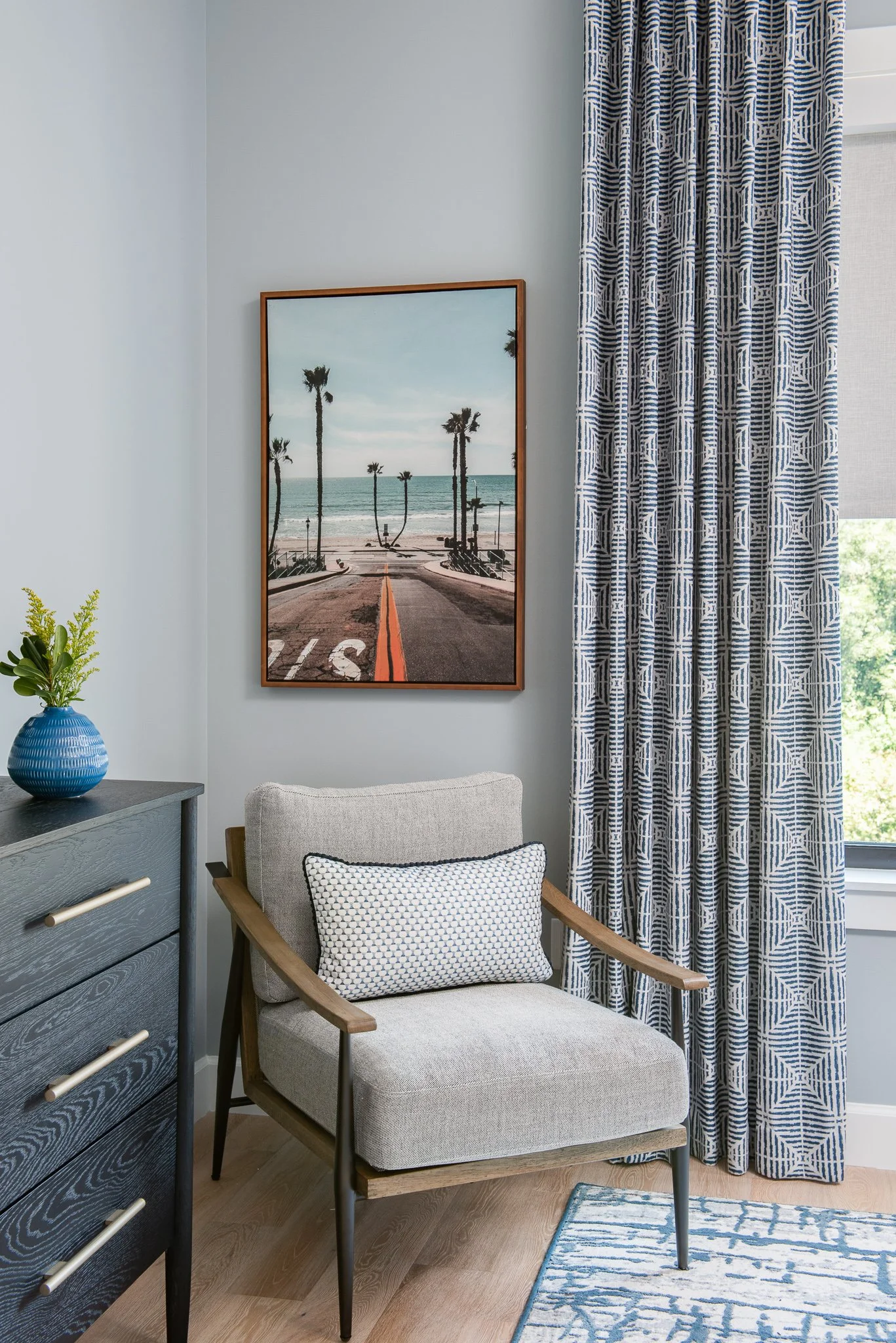 Interior of a room with a gray armchair with a patterned pillow, a blue dresser with a green plant in a blue vase, a framed picture of a beach scene with palm trees, patterned curtains, and a window revealing greenery outside.